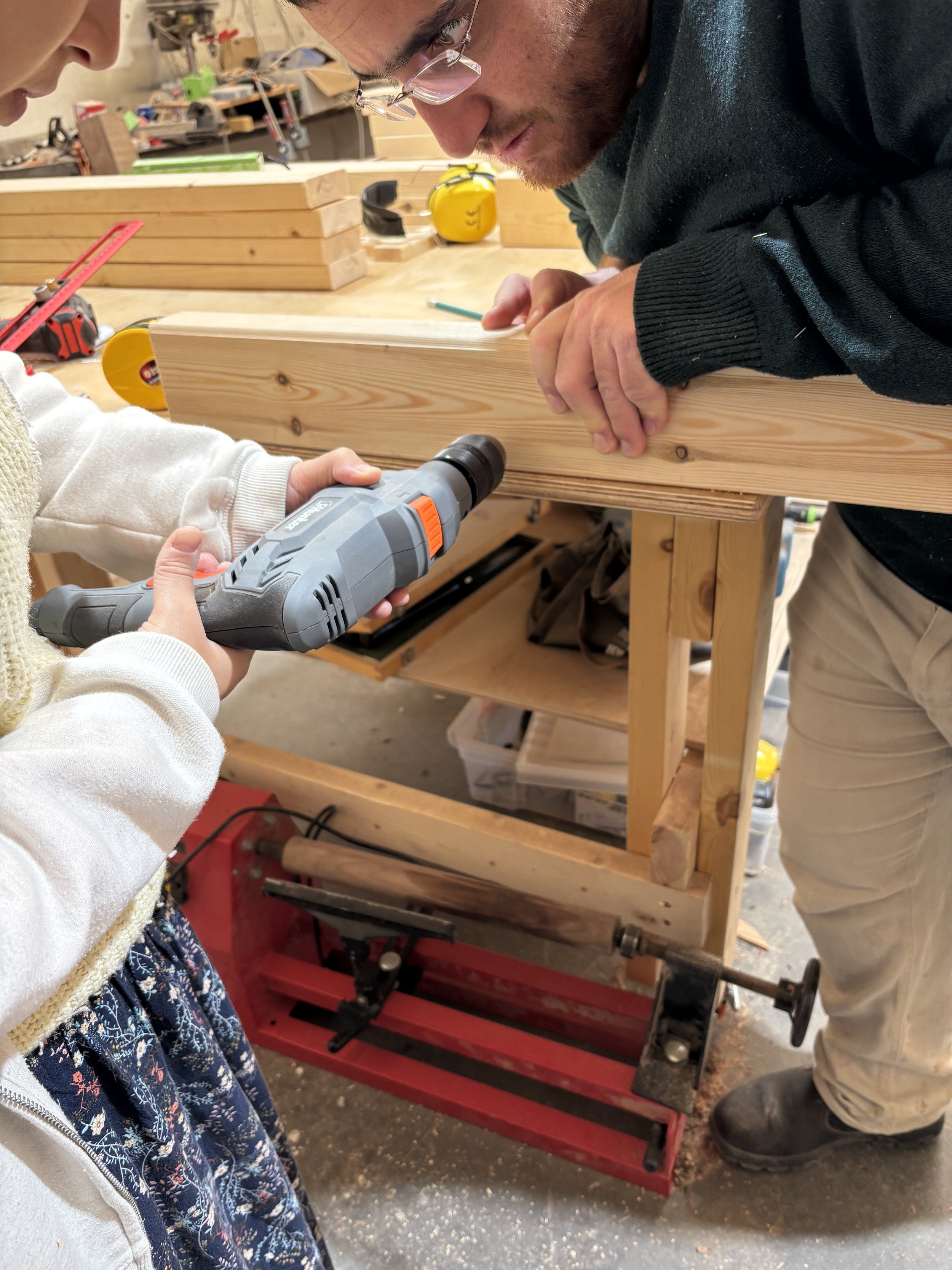 A person and a child working together in a woodworking shop, with the child holding a drill and the adult holding a piece of wood, while they assemble a piece of furniture or a woodworking project.