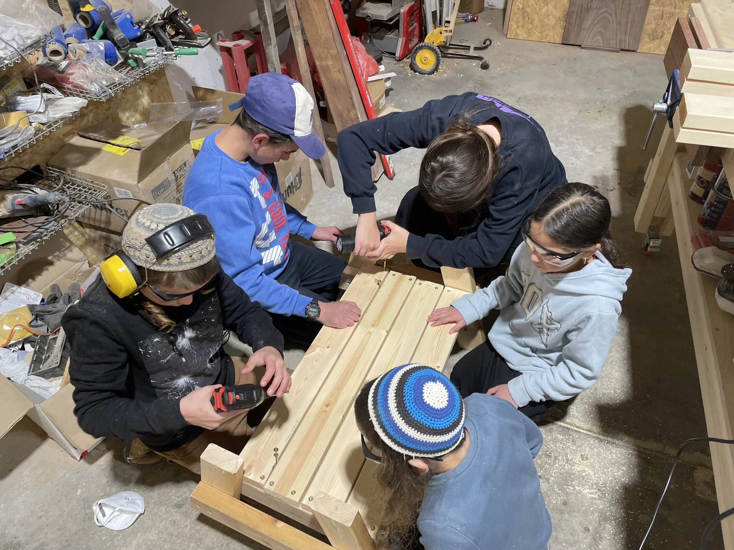 Group of five children working together on a woodworking project in a workshop. They are assembling a wooden piece, with tools and supplies around them.