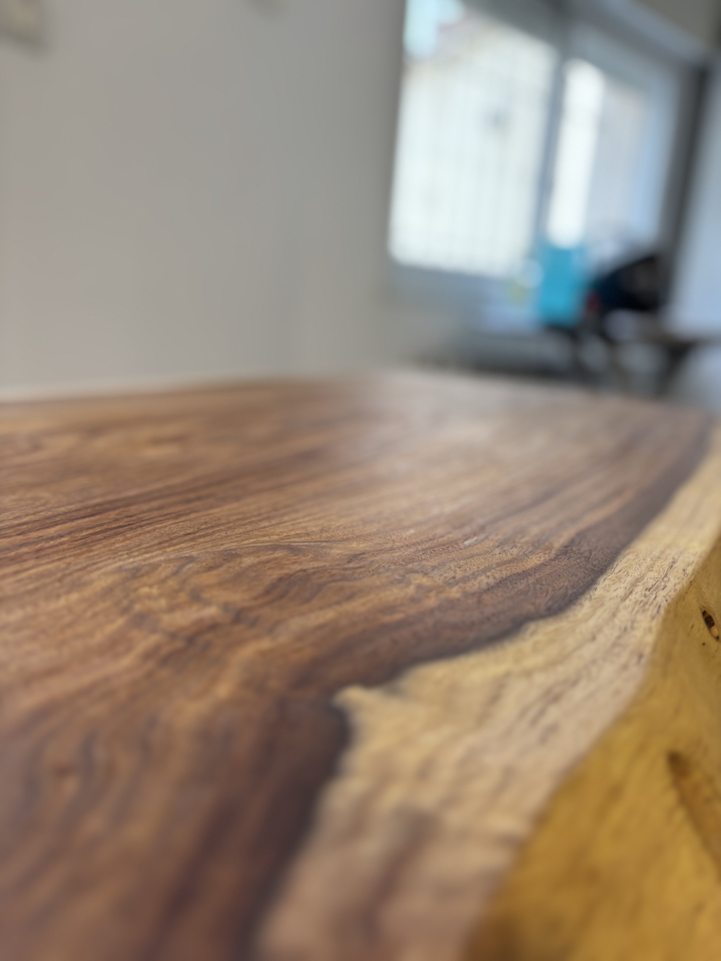 Close-up of a wooden surface with visible grain and texture, with a blurred background showing a window and some indoor objects.