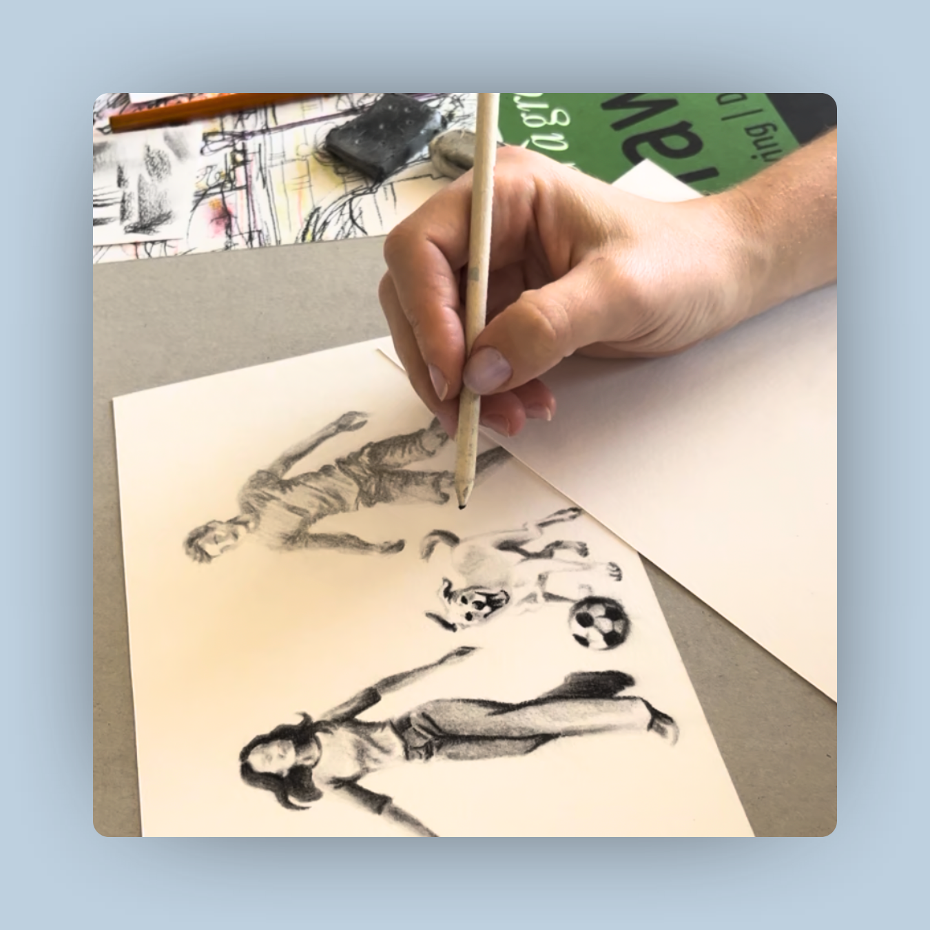 Muse&Flow Miami Art Commission Artist Patricia Machin book an artist Person drawing a sketch of a woman playing soccer with a dog and a ball on a piece of paper, with art supplies and sketchbooks in the background.