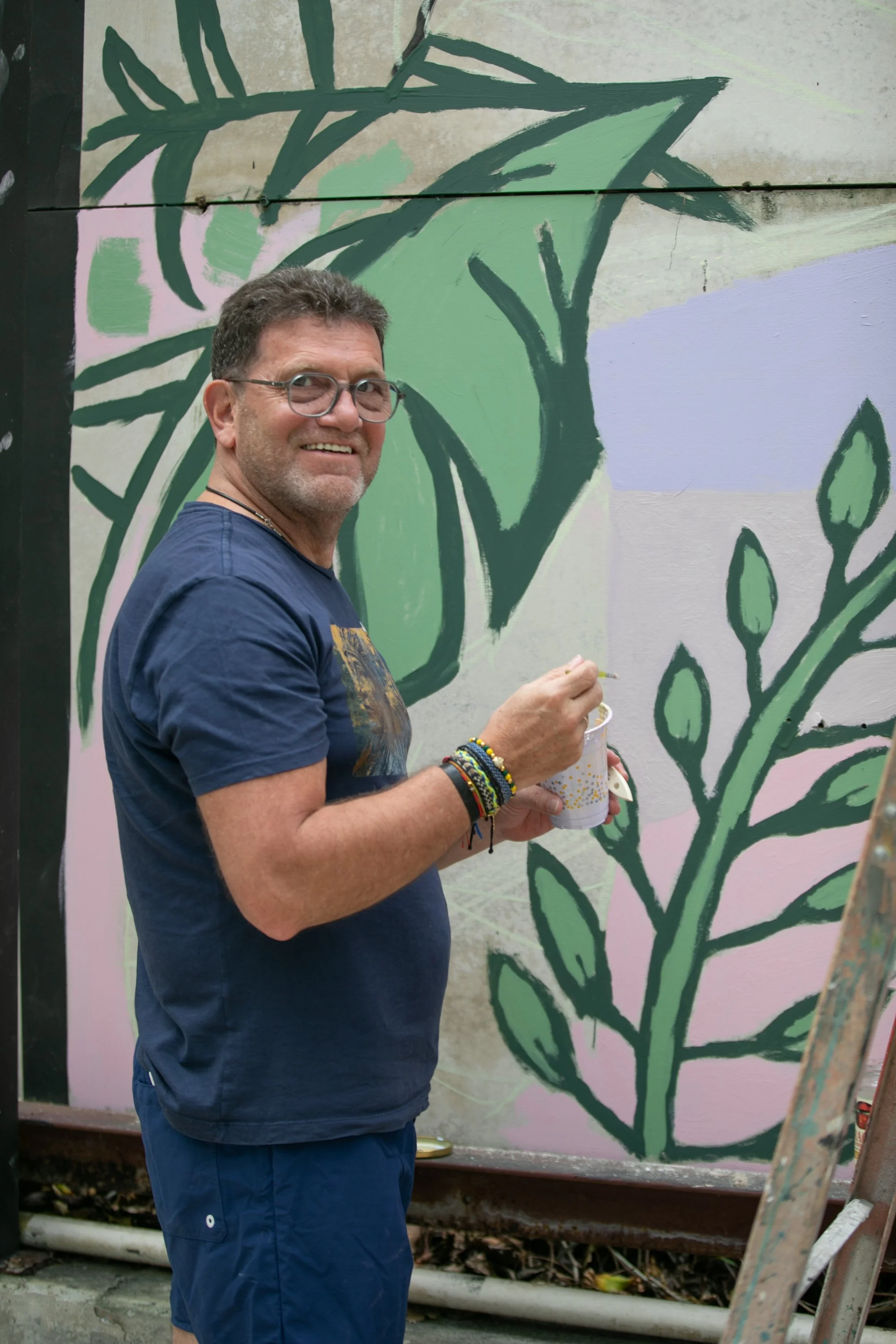 Muse&Flow Miami group custom handmade mural best ideas creative activities corporate team building art workshop Smile Human Ironside  A man with glasses and a beard smiling while holding a cup in front of a mural of green leaves painted on a wall.