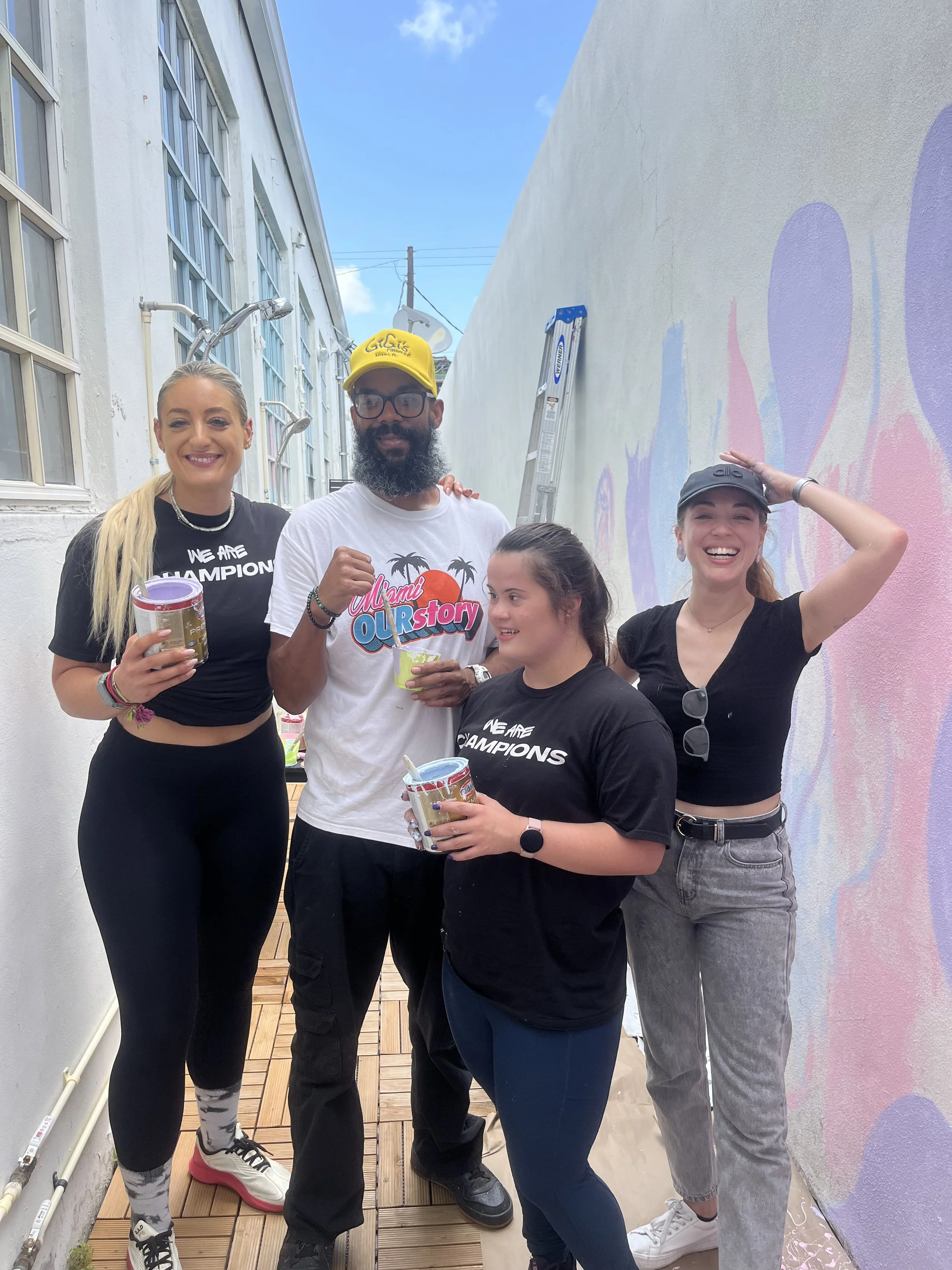 Muse&Flow Miami group custom handmade mural best ideas creative activities event activation team building art workshop Gigi Four women one man smiling and posing on a balcony, holding cups of paint, with a painted wall and ladder in the background.