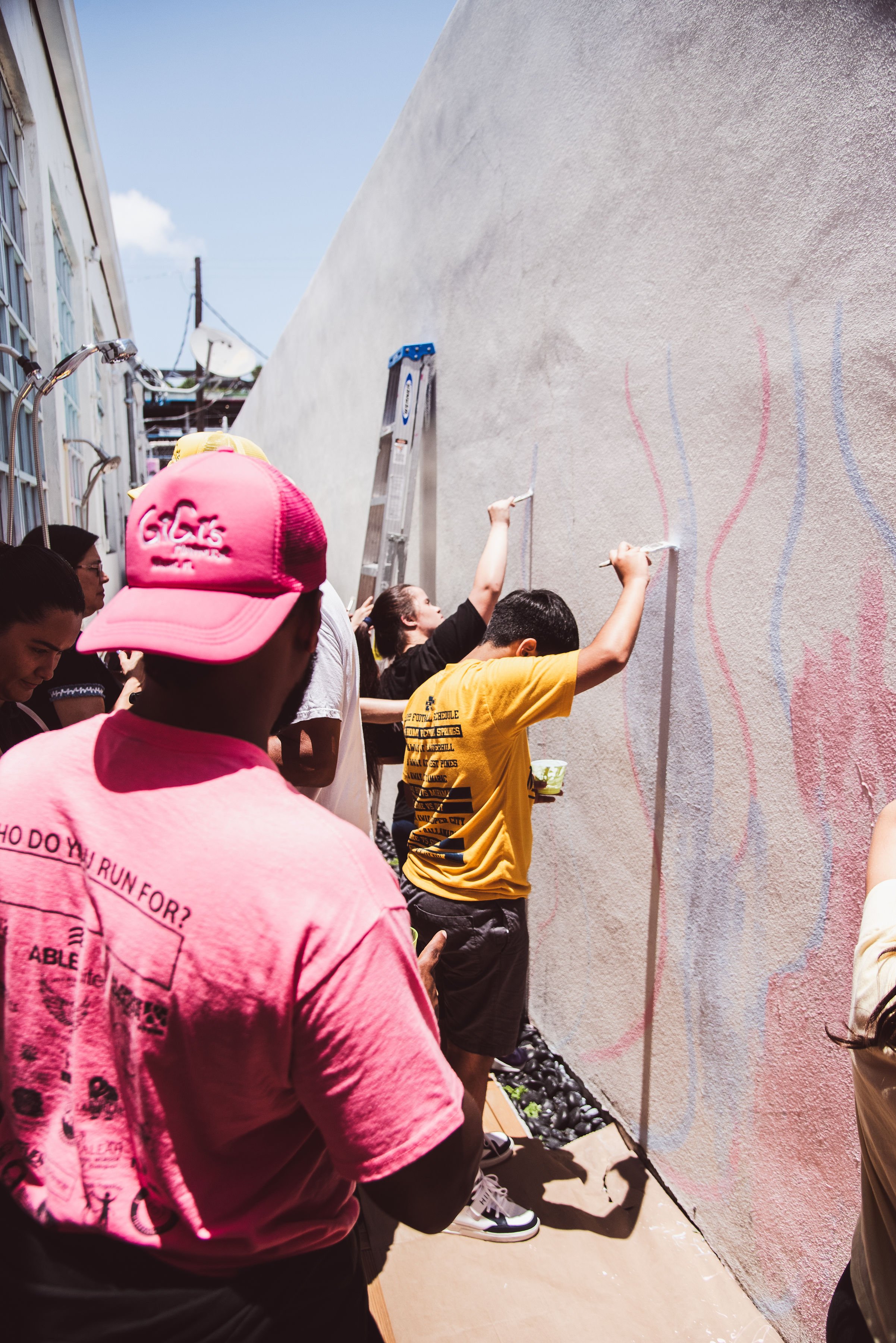 Muse&Flow Miami group custom handmade mural best ideas creative activities corporate team building art Group of people painting a mural on a outdoor wall, some are using brushes while others watch, with a ladder and balloons at the base of the wall.