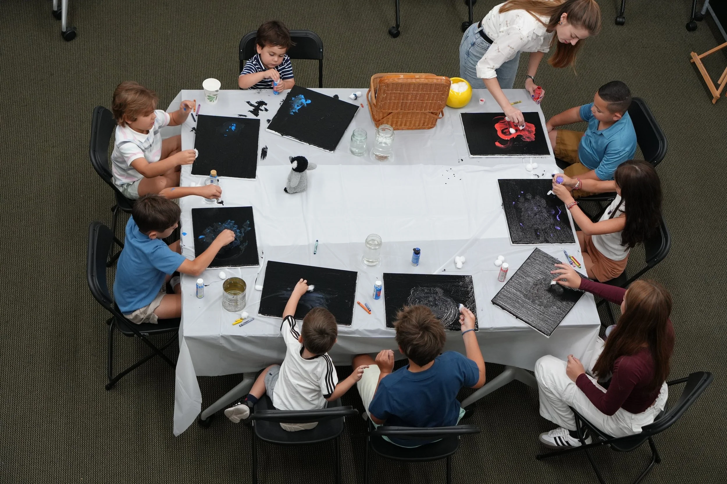 A group of children and an adult girl sitting around a rectangular table engaging in arts and crafts activities, creating pictures of animals on black canvases with paint and cotton balls.