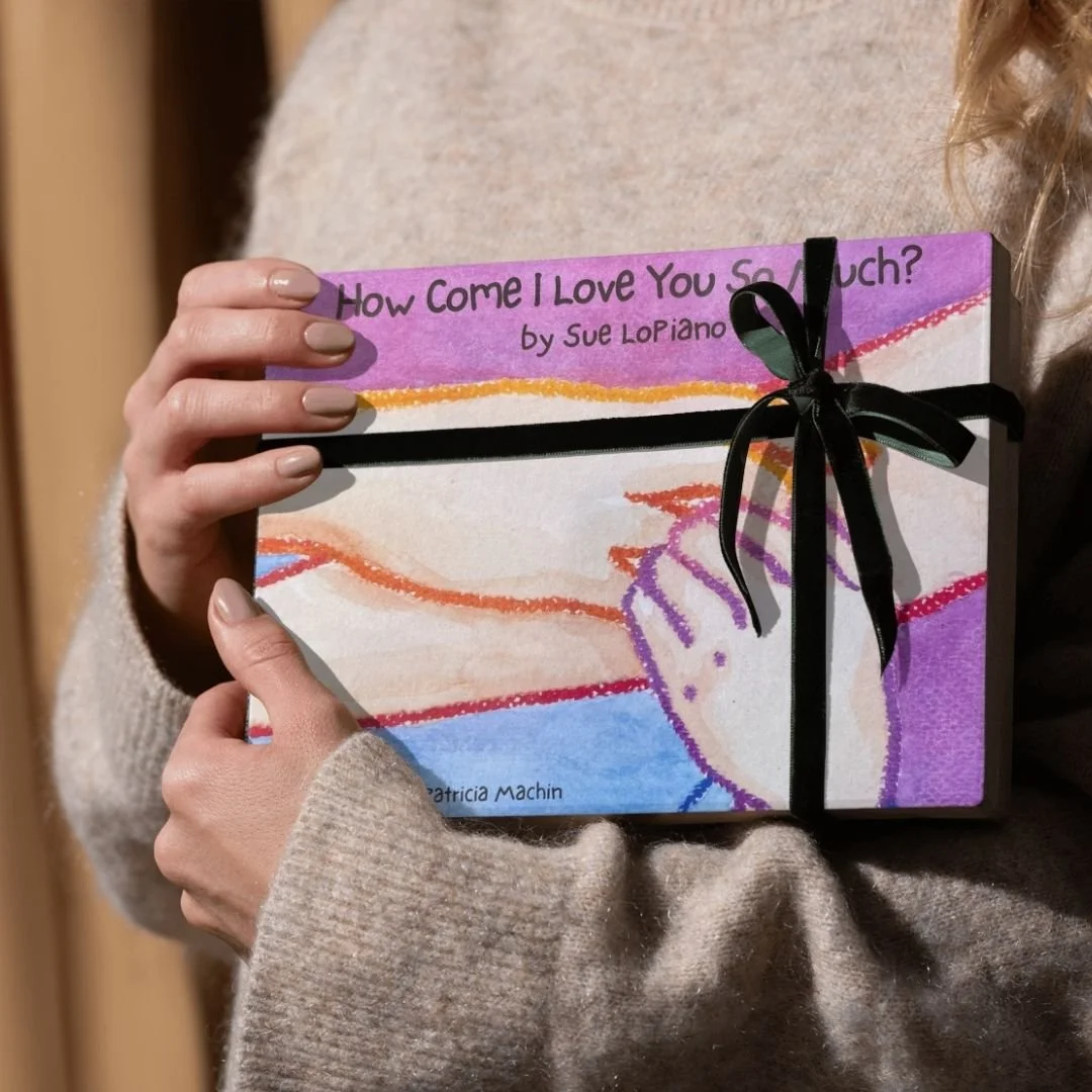 Muse&Flow Miami Art Services Commission Artist Patricia Machin book an artist A person in a beige sweater holding a colorful book titled "How Come I Love You So Much?" by Sue Lopianno, decorated with a black ribbon bow.