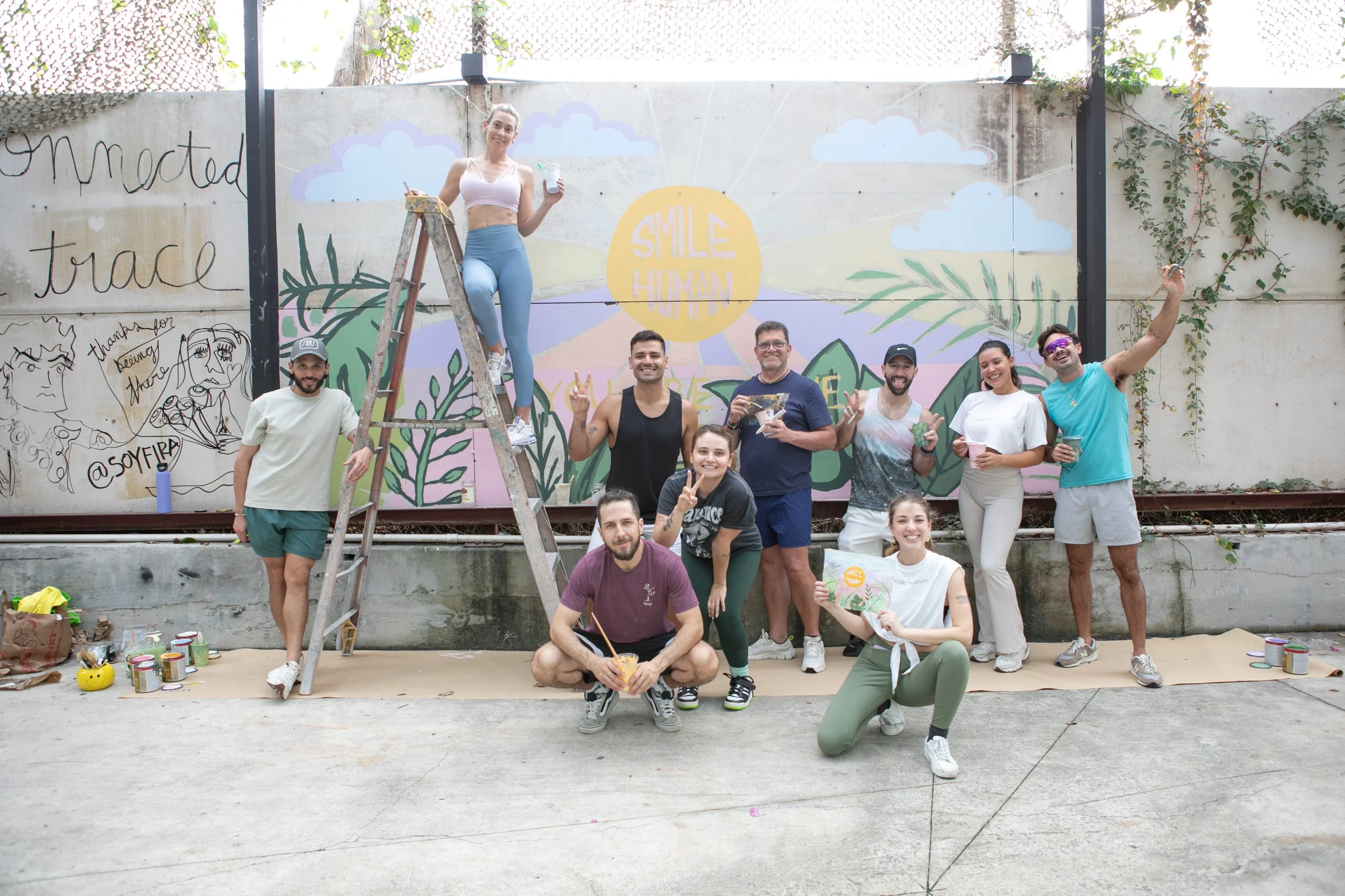 Muse&Flow Miami group custom handmade mural best ideas people participating in outdoor painting, paintbrushes and cups, with a colorful mural featuring clouds, plants, and the phrase "Smile Human" in the background. ironside