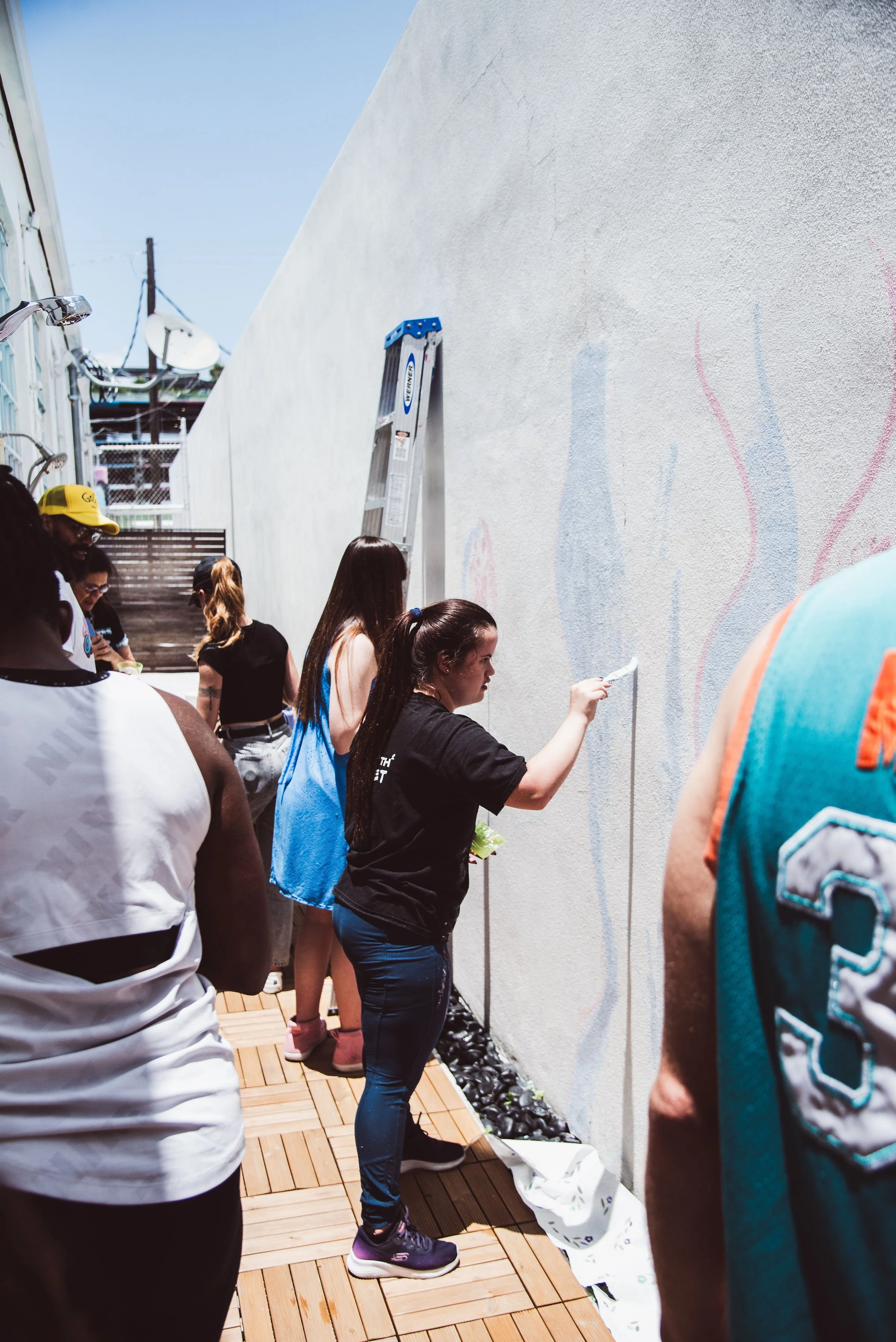 Muse&Flow Miami group custom handmade mural best ideas creative activities corporate team building art People gathered outdoors near a white wall, some painting or working on it, with a ladder and canopy in the background, and a wooden deck floor.
