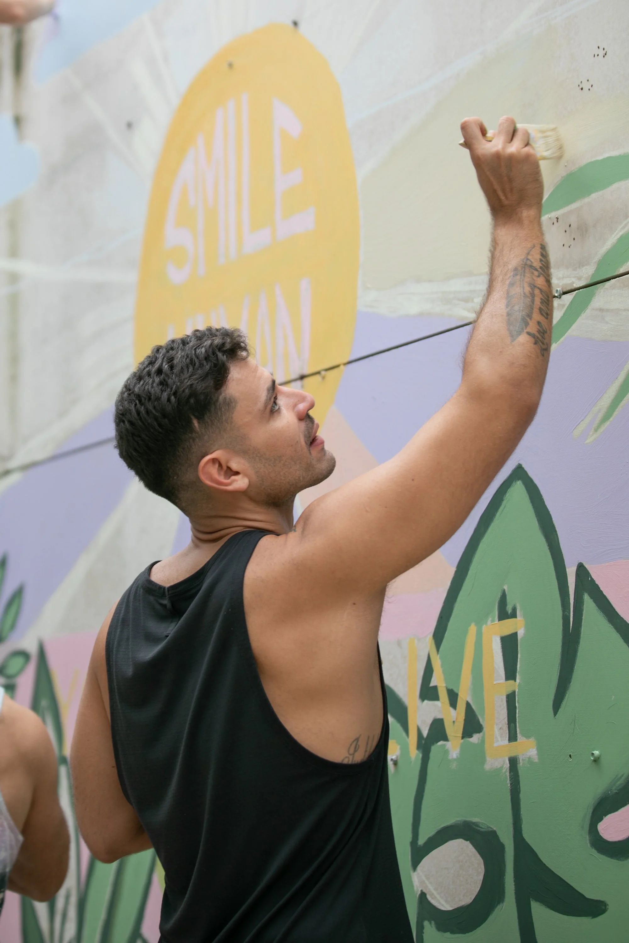 Muse&Flow Miami group custom handmade mural best ideas creative activities corporate team building art workshop Smile Human Ironside  A man with curly hair and tattoos on his arm painting a mural with pastel colors on a wall.