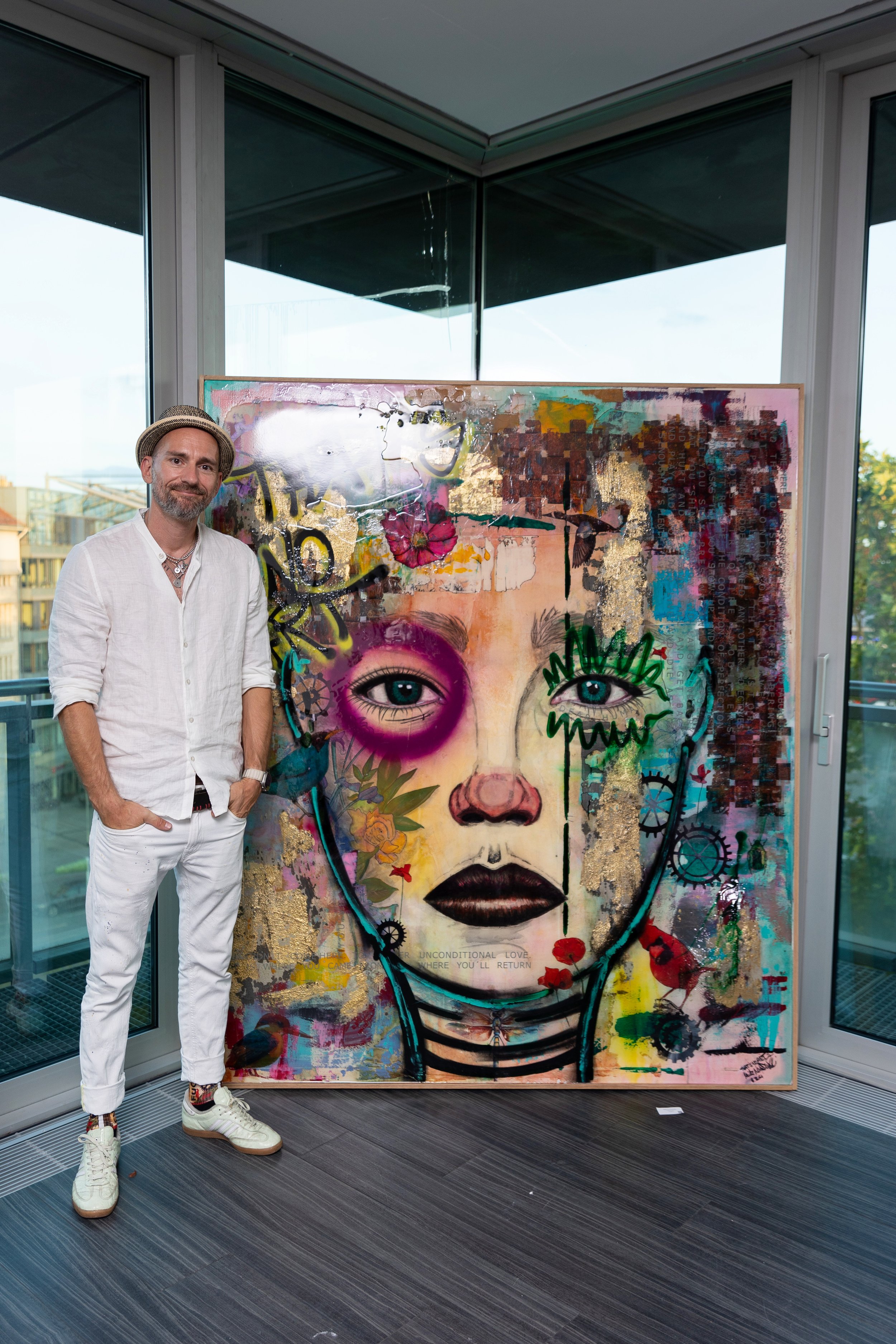 Ein Mann in weißem Outfit und Hut steht neben einem großen, bunten Kunstwerk eines Gesichtes mit mehreren Gesichtsdetails, Blumen, Graffiti-Elementen und goldenen Akzenten, innerhalb eines modernen Innenraums mit großen Fenstern.