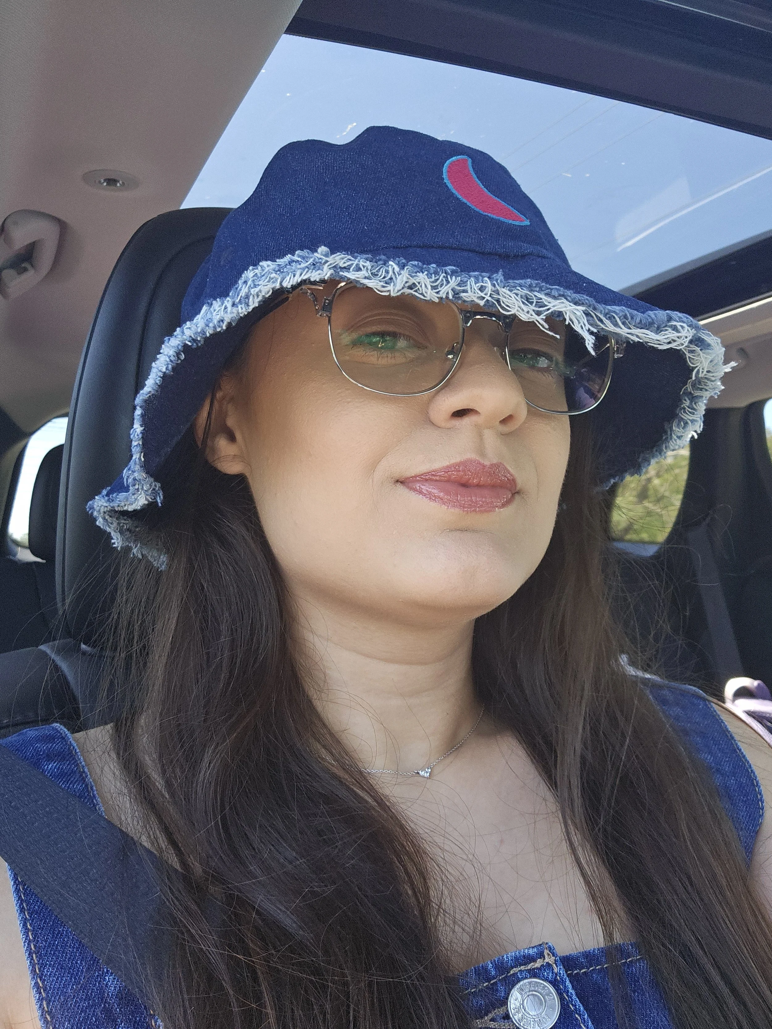 A woman with long brown hair wearing a blue denim hat with a red and blue logo, sunglasses, a blue denim jacket, and a silver necklace, sitting inside a vehicle with a sunroof, looking out the window.