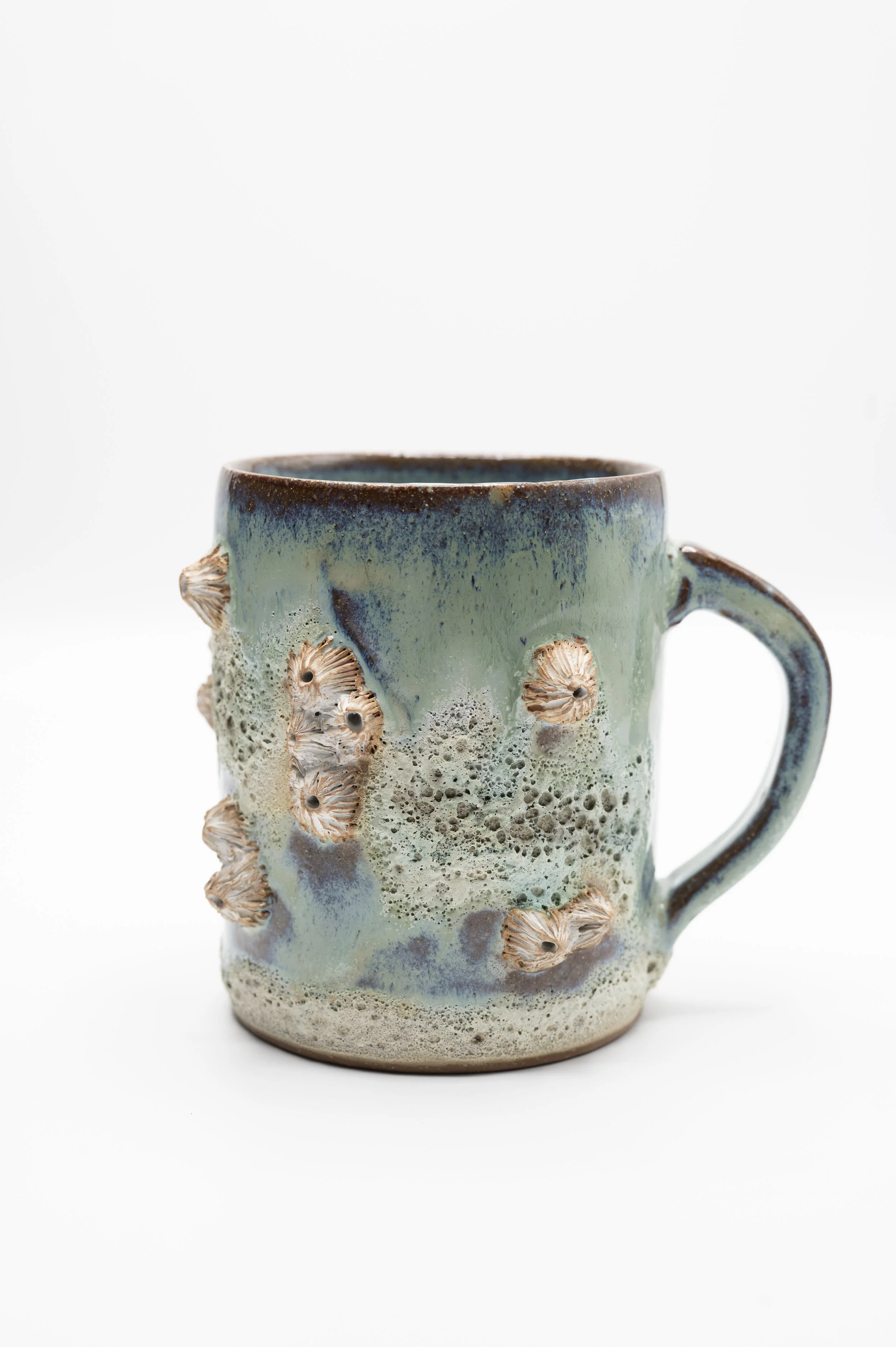 A ceramic mug with a textured surface, decorated with barnacles glazed with blue, green, brown, and speckled colors, and featuring a curved handle, set against a white background.