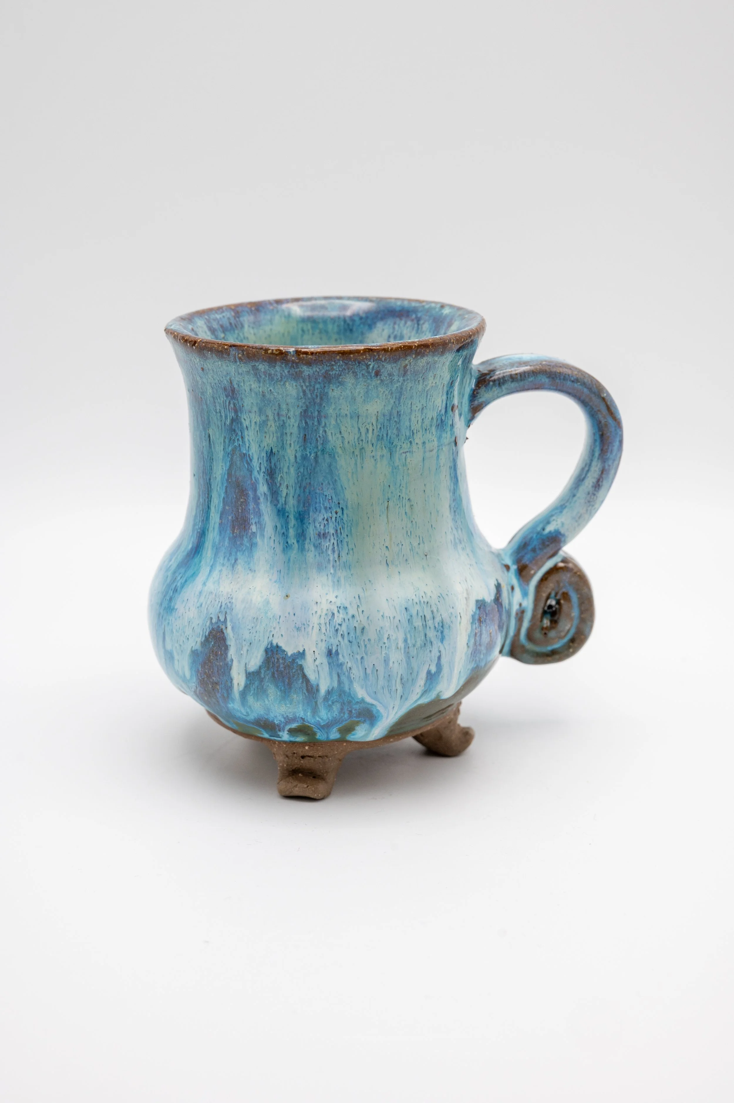 A ceramic mug with a blue and green glaze, featuring a round body, a curved handle, and three small feet, set against a plain white background.