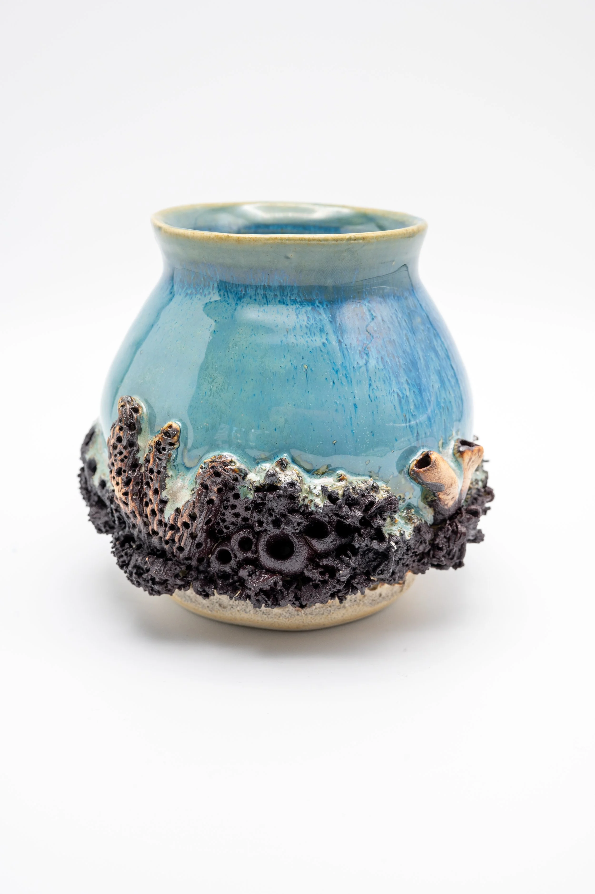 A ceramic vase with a blue and teal glazed top half and a textured dark brown, black, and tan carved base resembling coral or lava, against a plain white background.