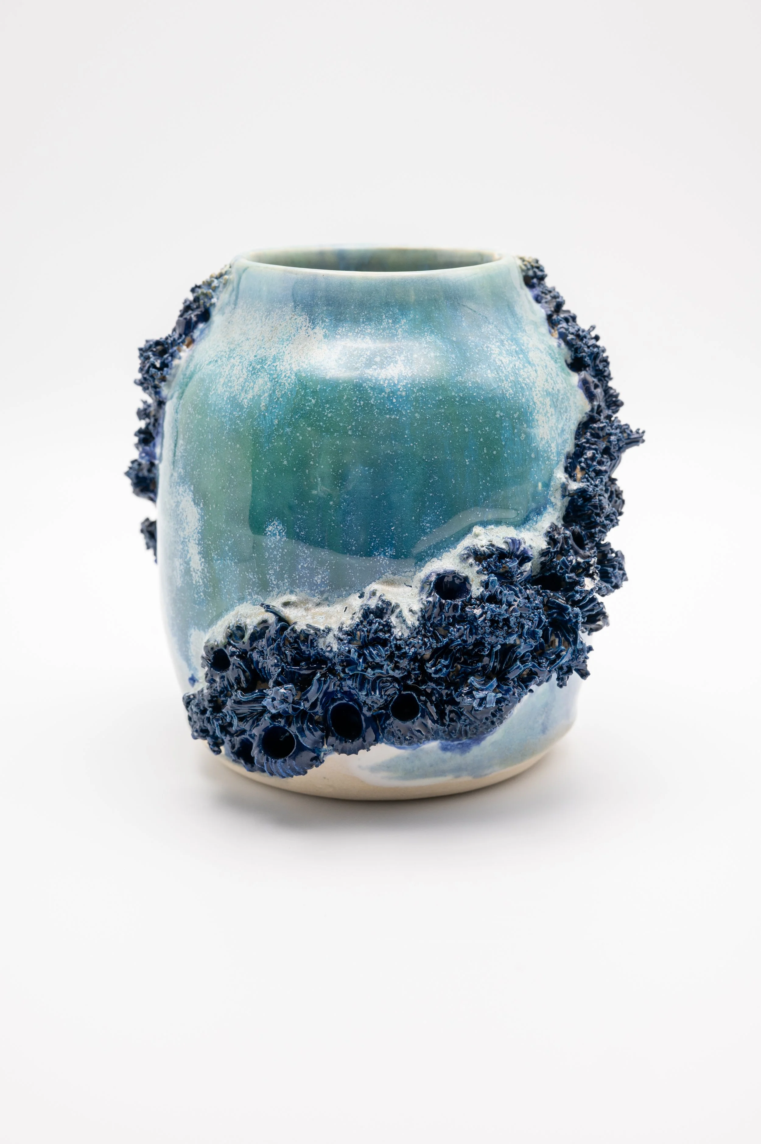 A ceramic vase with a smooth, glossy surface in shades of green and blue. The vase features a textured, dark blue, flower-like pattern along the sides, giving it an artistic and modern appearance.