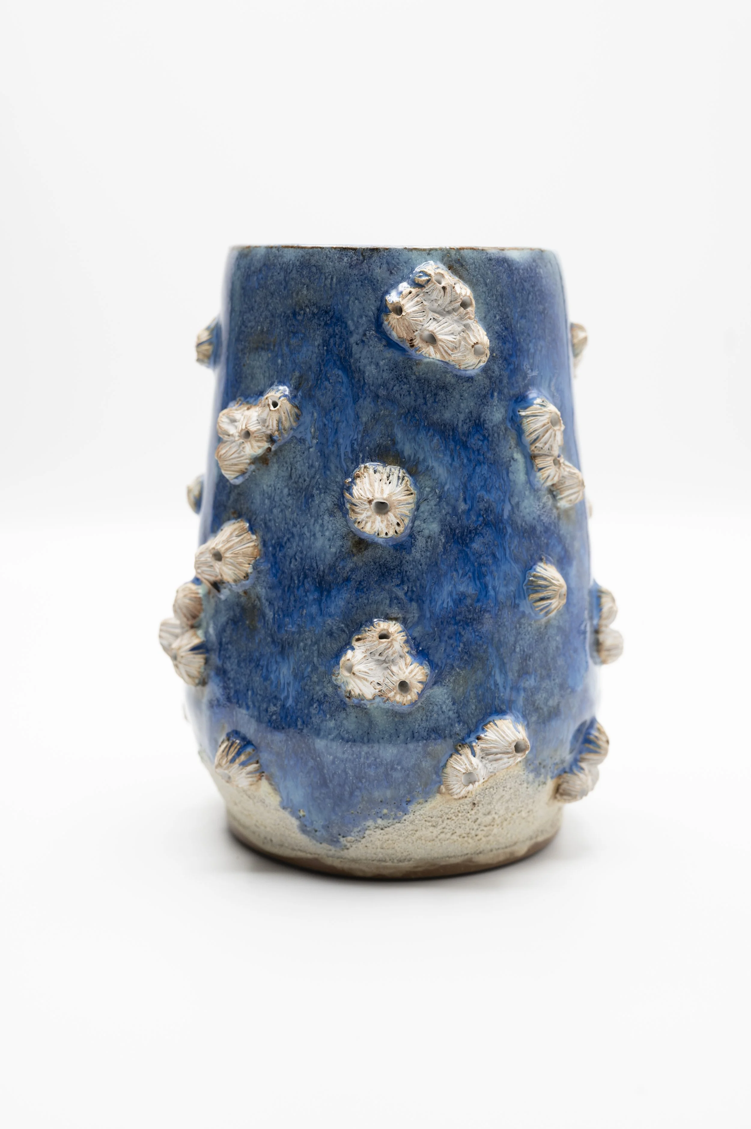 A ceramic vase with a blue glaze and decorated with small seashells protruding from its surface, set against a plain white background.