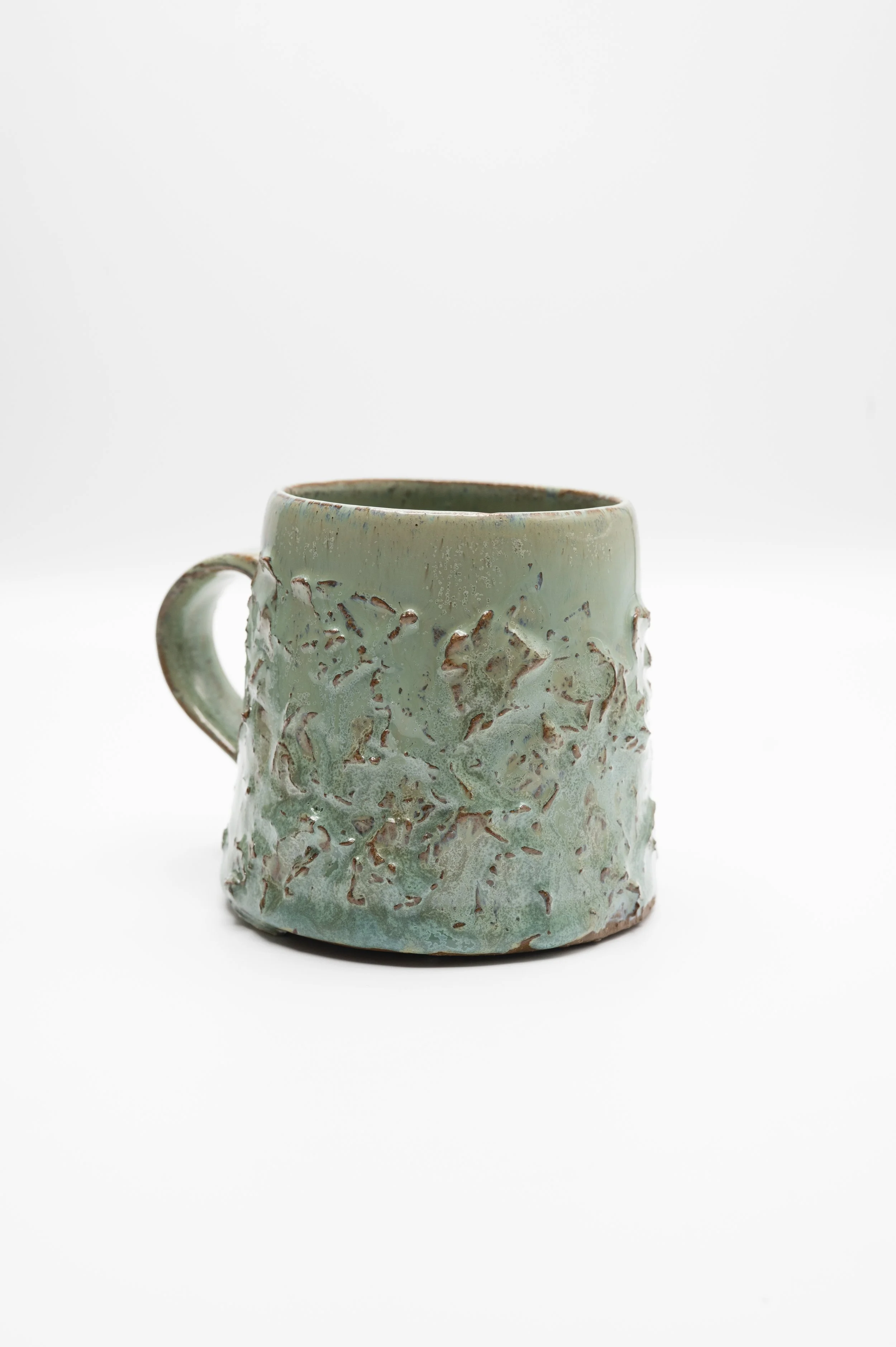 A ceramic mug with a textured, greenish surface and a handle, set against a plain white background.