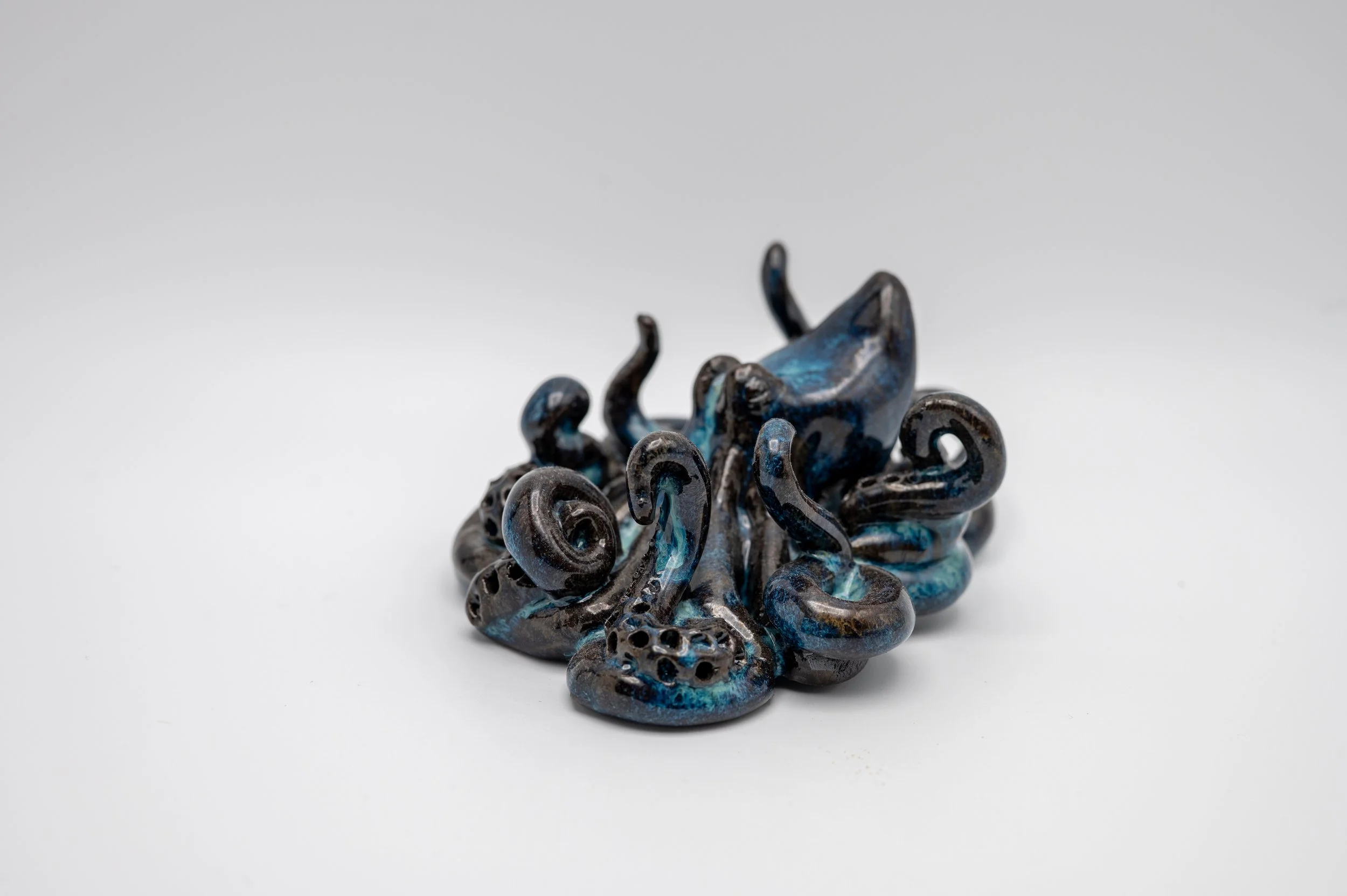 A ceramic sculpture resembling an octopus with curled tentacles, predominantly black with blue accents, set against a plain white background.
