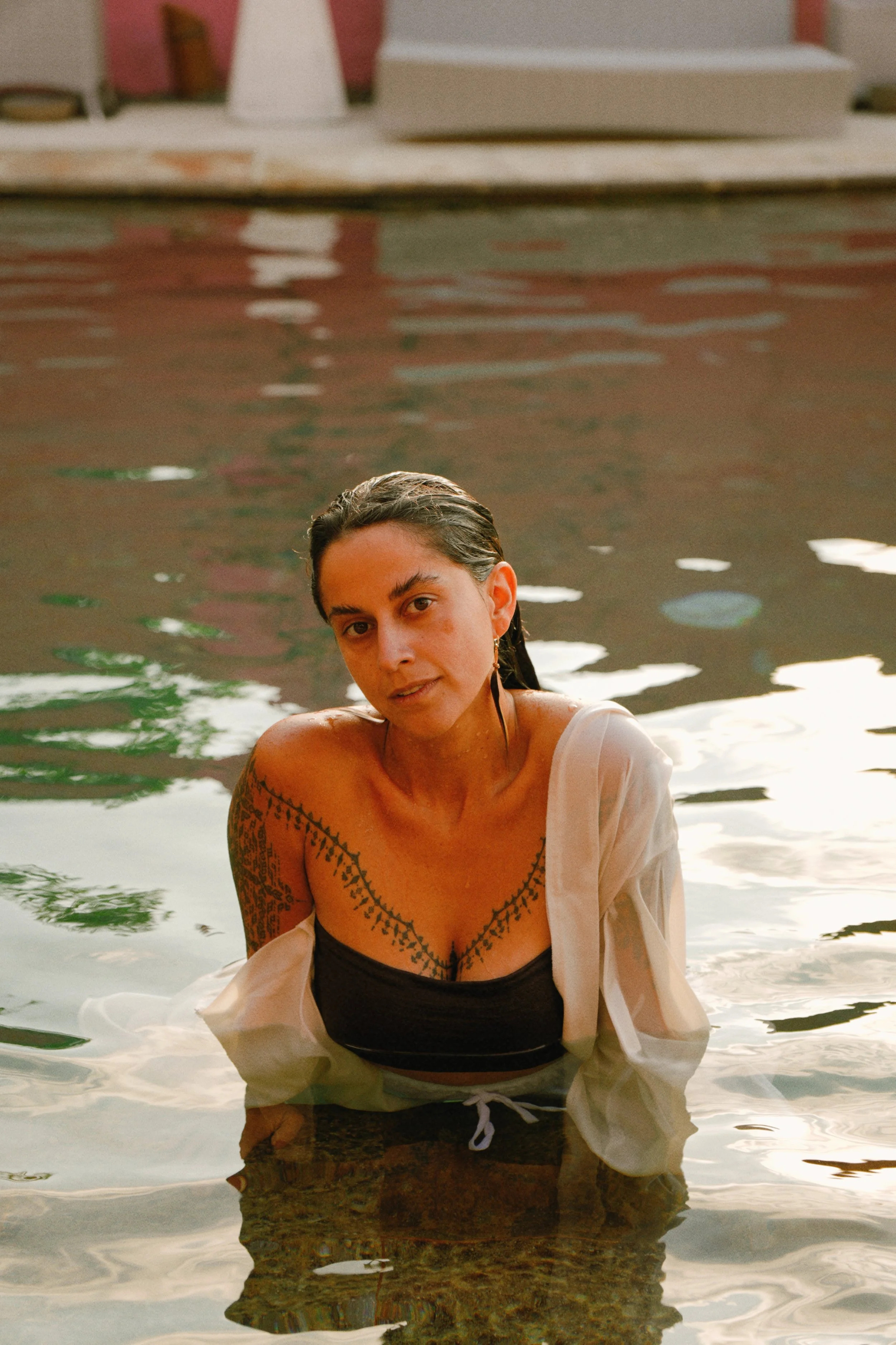 A woman with dark hair tied back, wearing a black top and white shirt, sitting in a body of water with a wooden dock and building in the background.