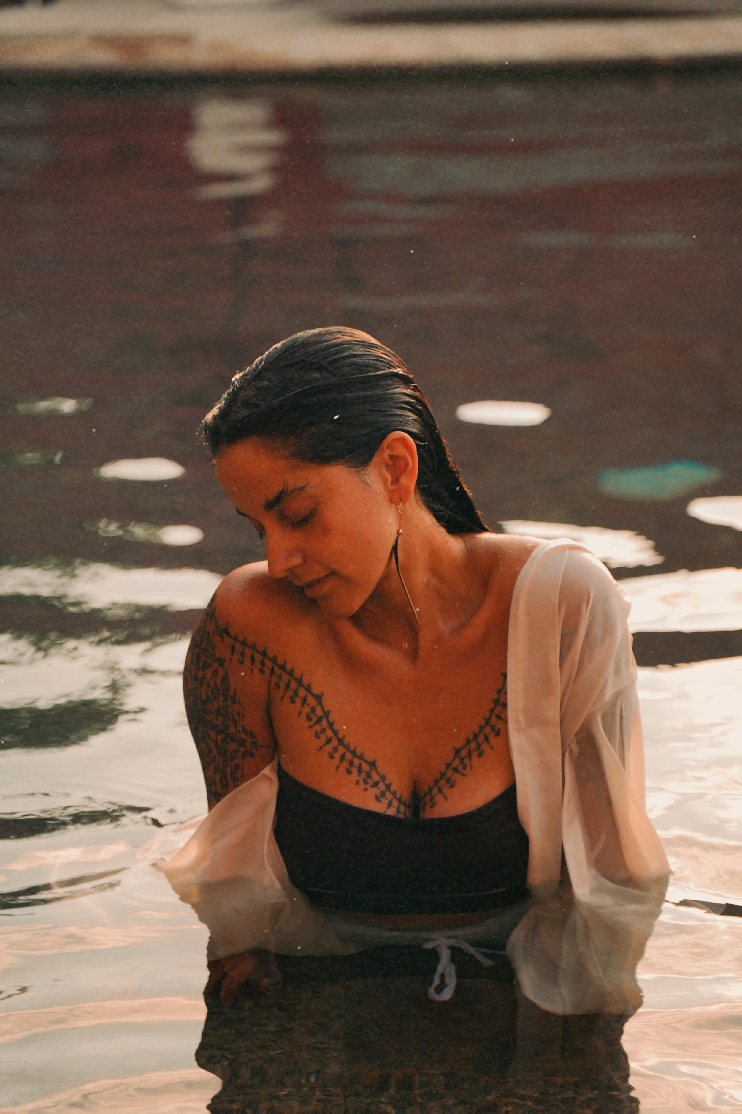 A woman with wet hair in a white shirt and black top, and tattoos on her arm, is sitting in water with her head tilted down and eyes closed during sunset.