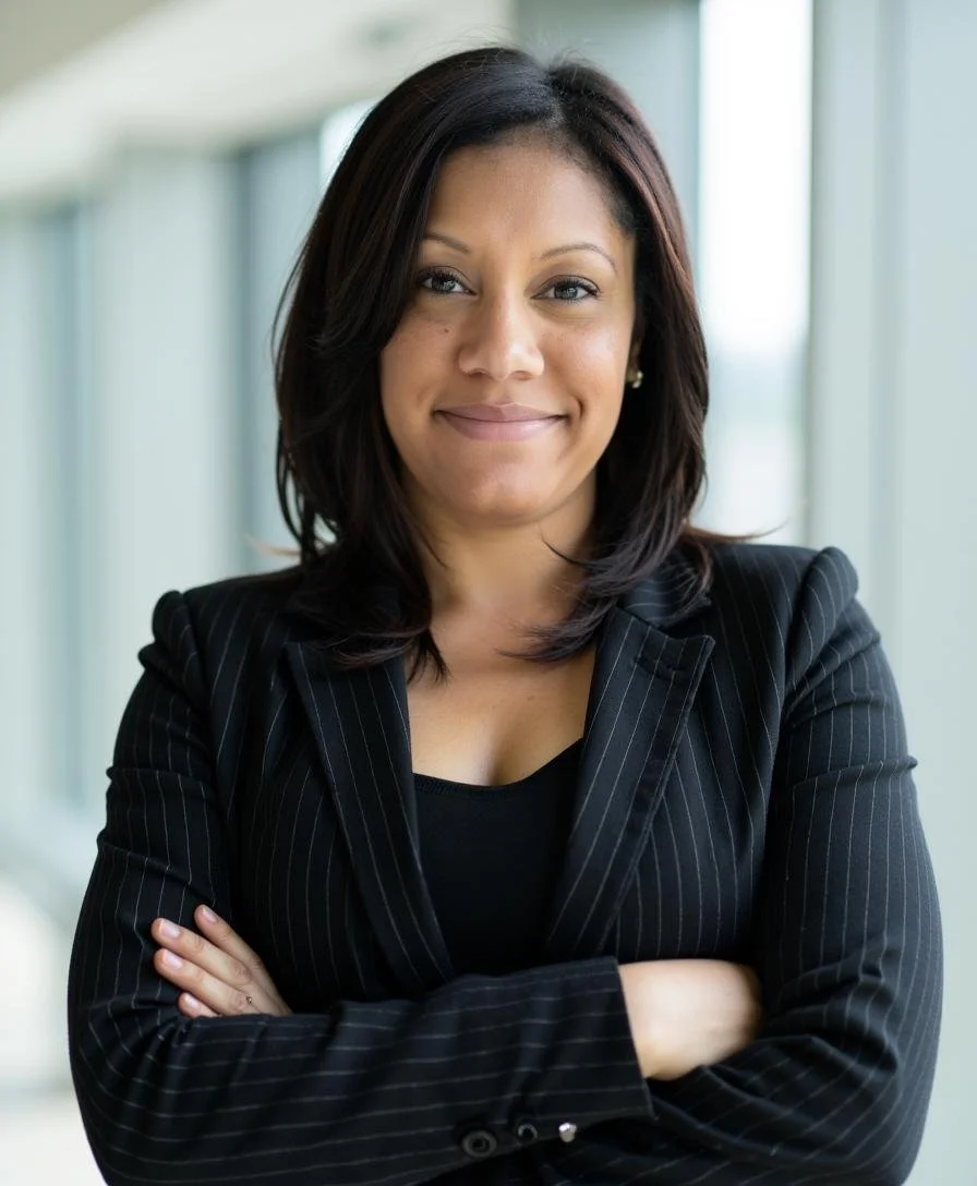 Woman in a black pinstripe blazer with arms crossed, smiling, standing in a bright indoor setting.