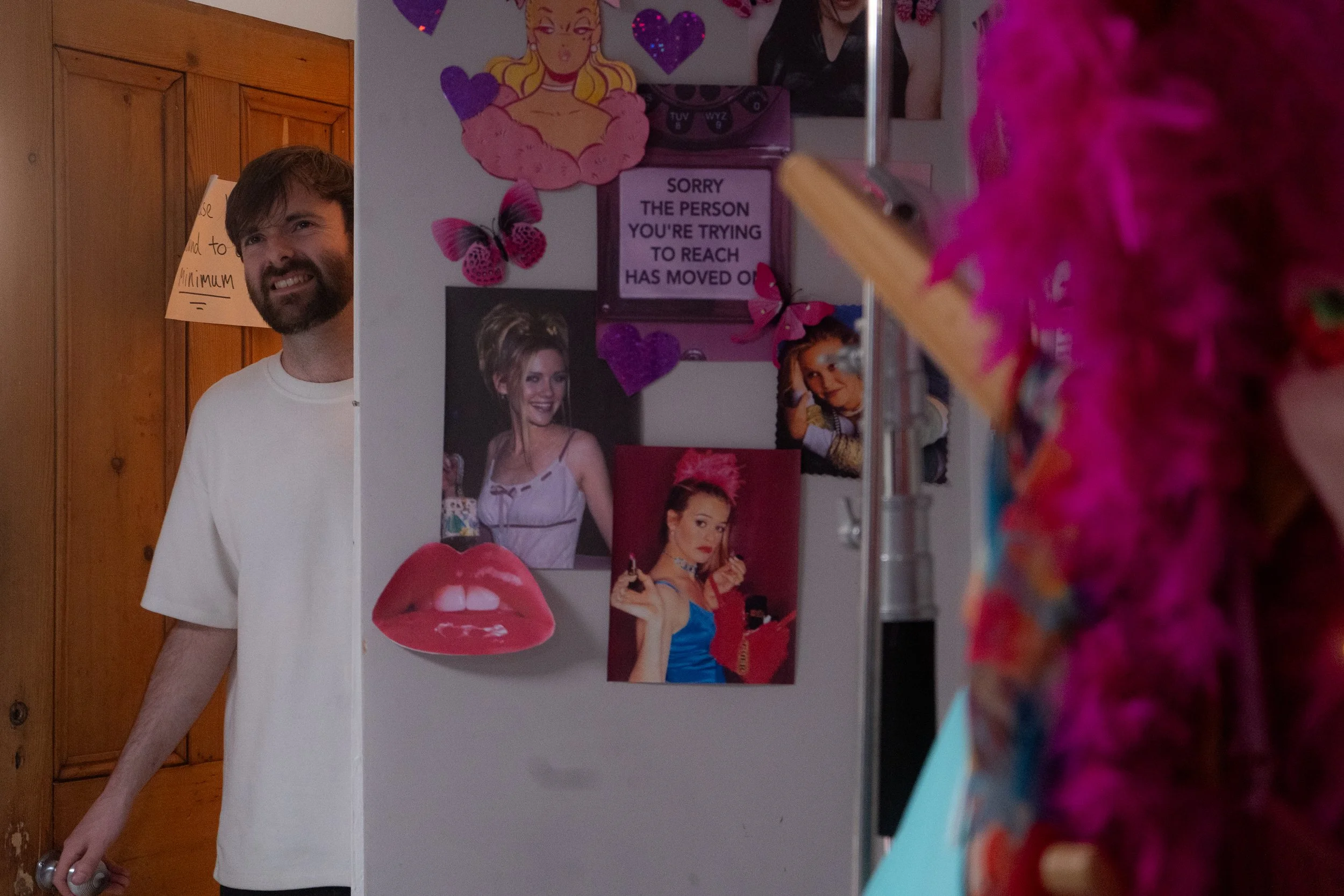 A man with a beard and white T-shirt smiling as he looks around a doorframe into a room. The room's wall is decorated with various colorful posters, photos, and cutouts, including a sign that reads 'Sorry the person you're trying to reach has moved o