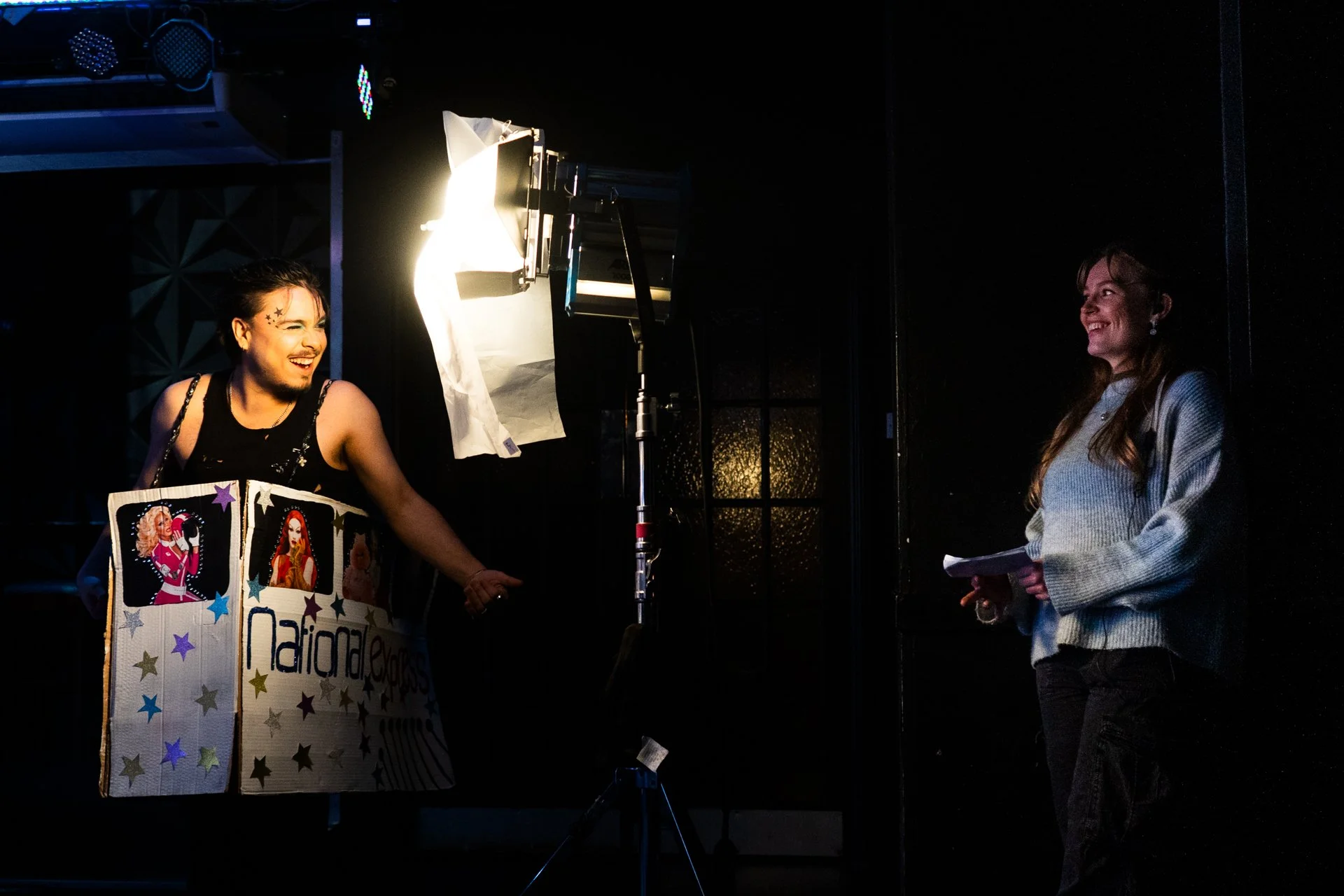 A person on stage wearing a black outfit and a large cardboard box costume with colorful stars and images of women, smiling and engaging with a woman holding a notepad, in a dark room with stage lighting.
