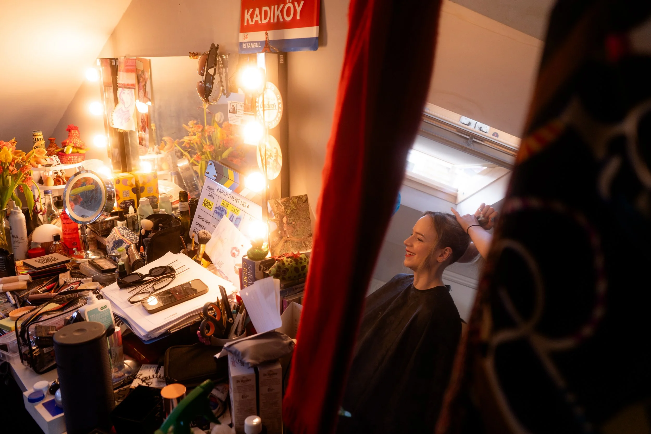 A woman smiling in a makeup chair, seen through red curtains, with a cluttered vanity table filled with makeup, skincare, and beauty tools, illuminated by bright lights.