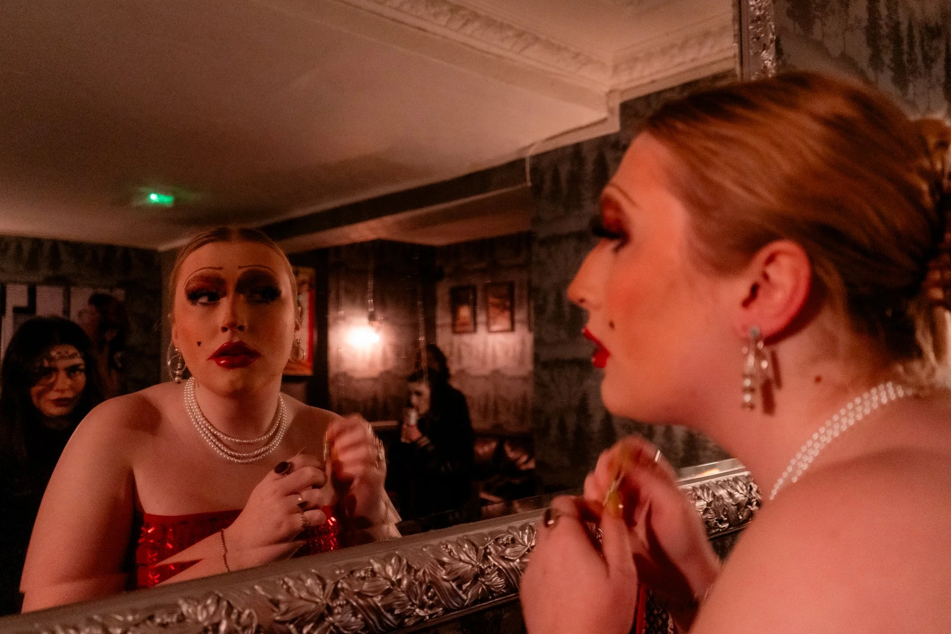 A person with dramatic makeup, red lipstick, and jewelry looks at themselves in a mirror while holding a makeup brush. In the background, other people are visible in a dimly lit room with dark walls and vintage-style decor.