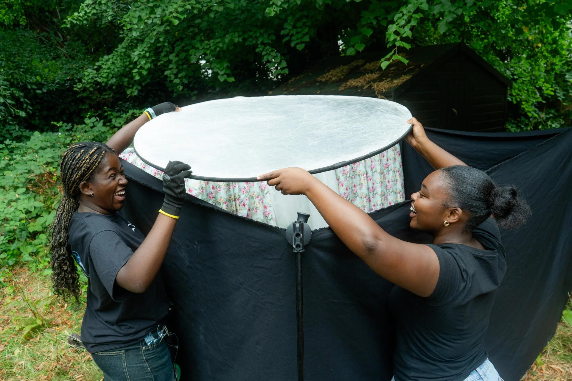 Two women adjusting a large round reflector outdoors surrounded by greenery, both smiling and wearing black shirts.