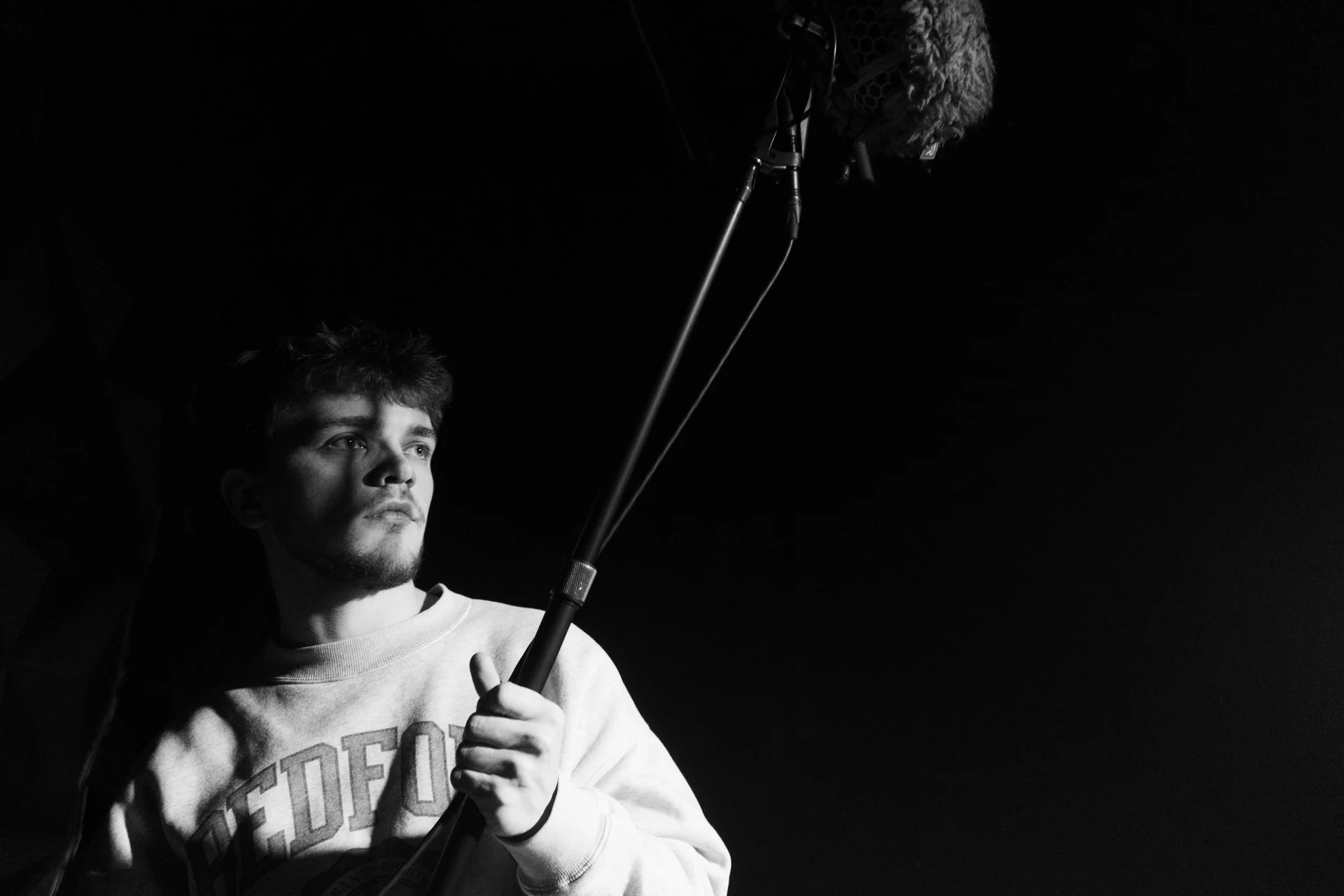 A young man holding a microphone boom pole, looking seriously at something off-camera, in a black-and-white setting with a dark background.