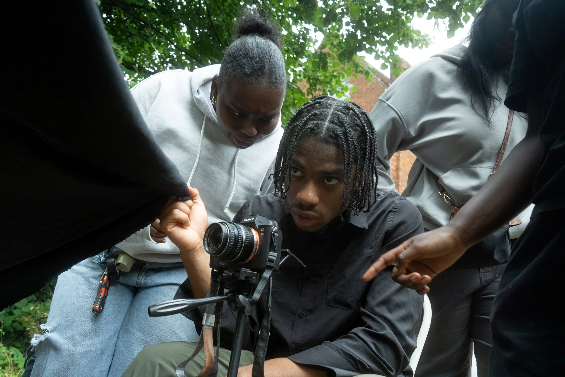 Group of four people gathered around a camera outdoors, looking at the viewfinder with focused expressions.