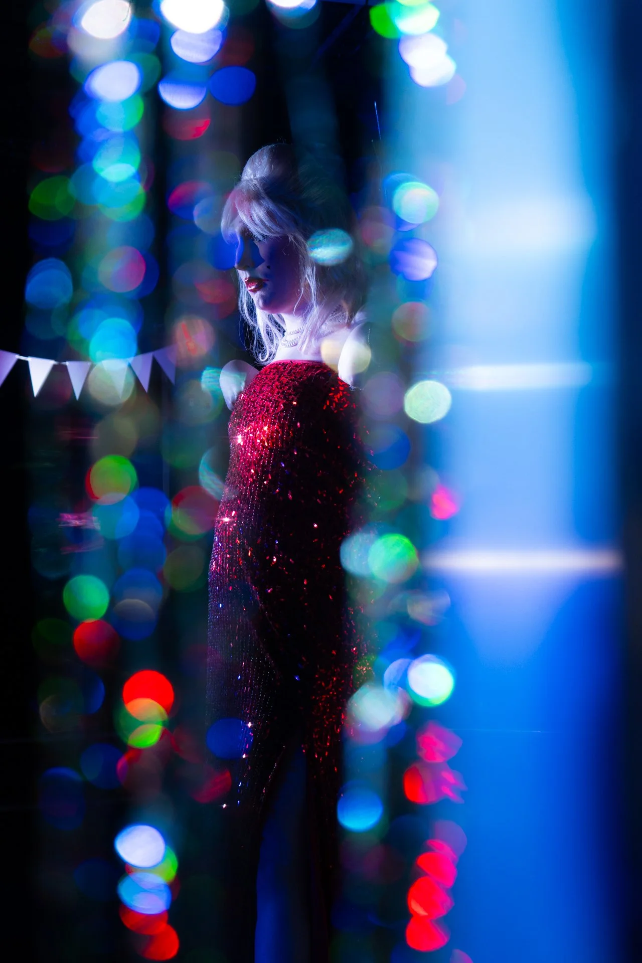 A woman in a red sequin dress standing behind a curtain of colorful bokeh lights, with a soft blue light illuminating her face from the right side.