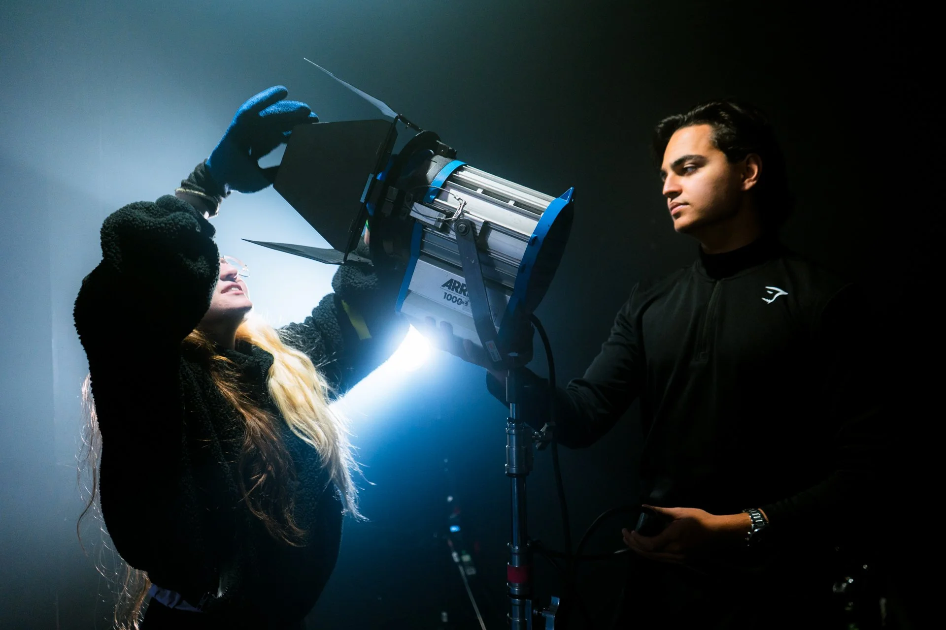 Two people setting up film or photography equipment in a dimly lit studio, with one person adjusting a light and the other watching.