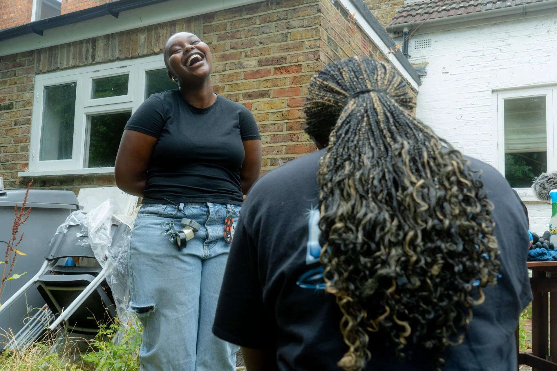 Two women are smiling and laughing outside by a brick house. One woman with short hair is standing, wearing a black t-shirt and ripped jeans. The other woman, with long braided hair, is sitting with her back to the camera.