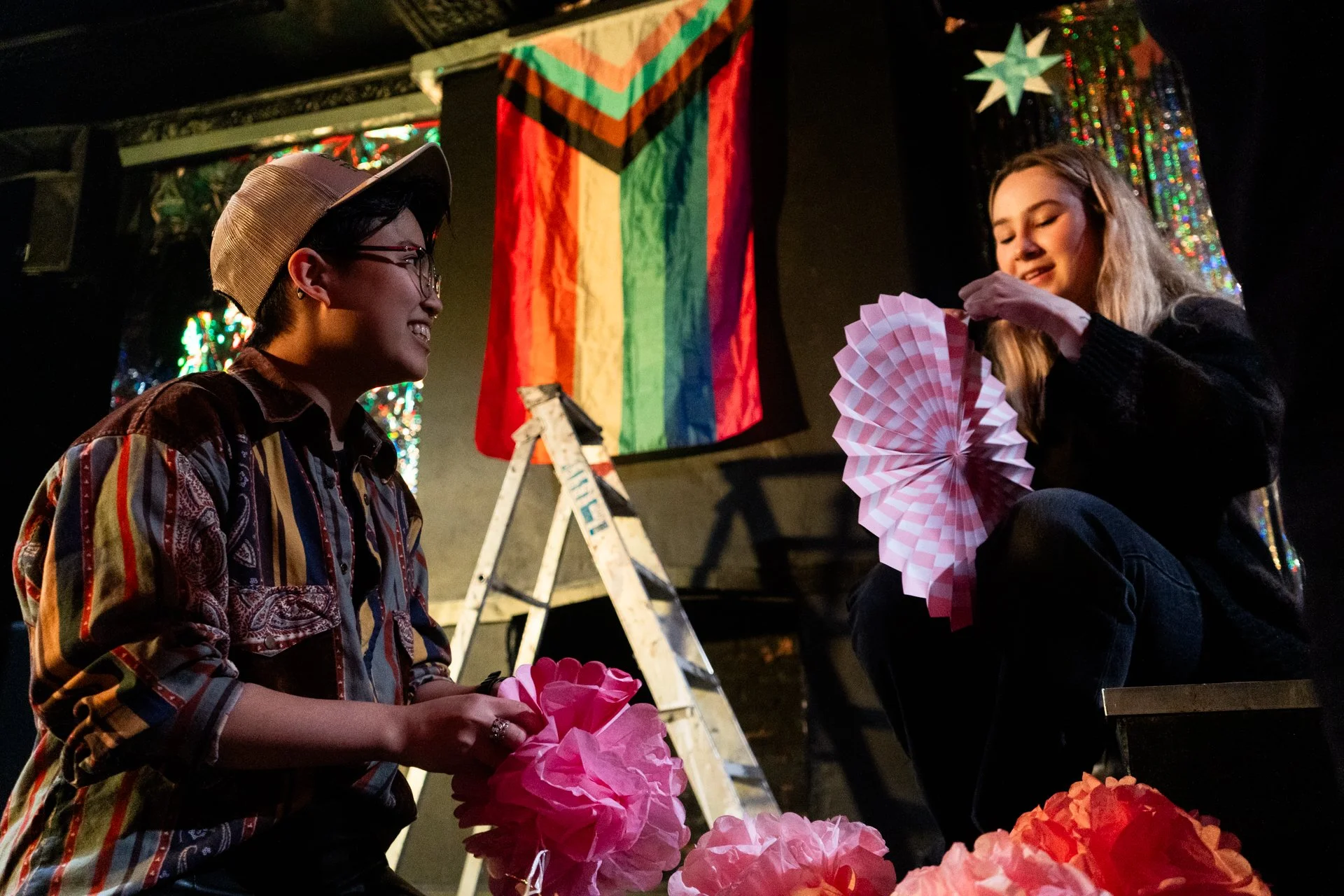 Two people decorating for Pride with rainbow flags and pink paper decorations.