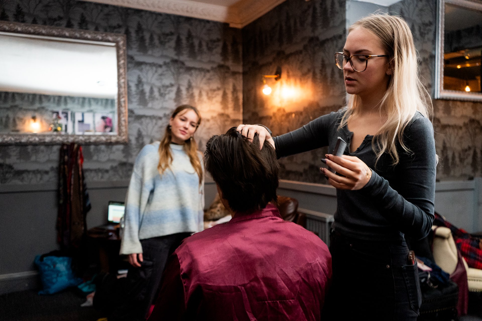 A blonde woman with glasses is styling a woman's hair in a salon, while another woman with long hair and a colorful sweater looks on.