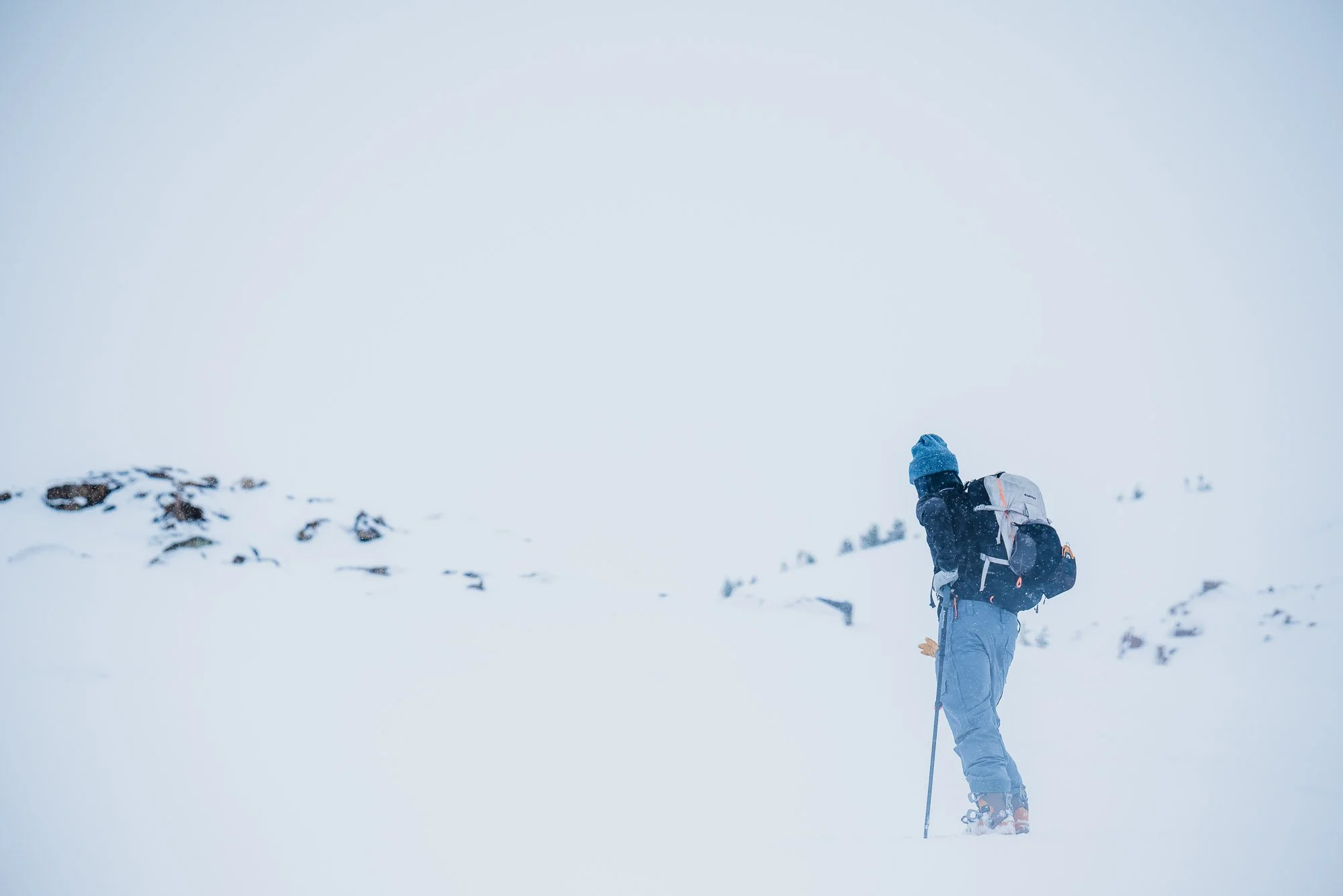 A person in winter gear, including a blue hat and jacket, hiking through a snow-covered landscape with snow and rocks visible in the background. Montana backcountry skiing