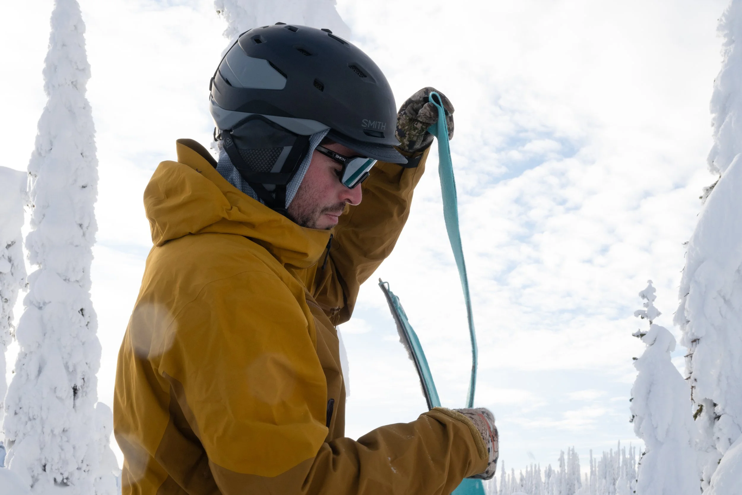 A man in a yellow jacket, wearing a black helmet, sunglasses, and gloves, is holding a blue strap in a snowy forest with snow-covered trees and sky in the background.