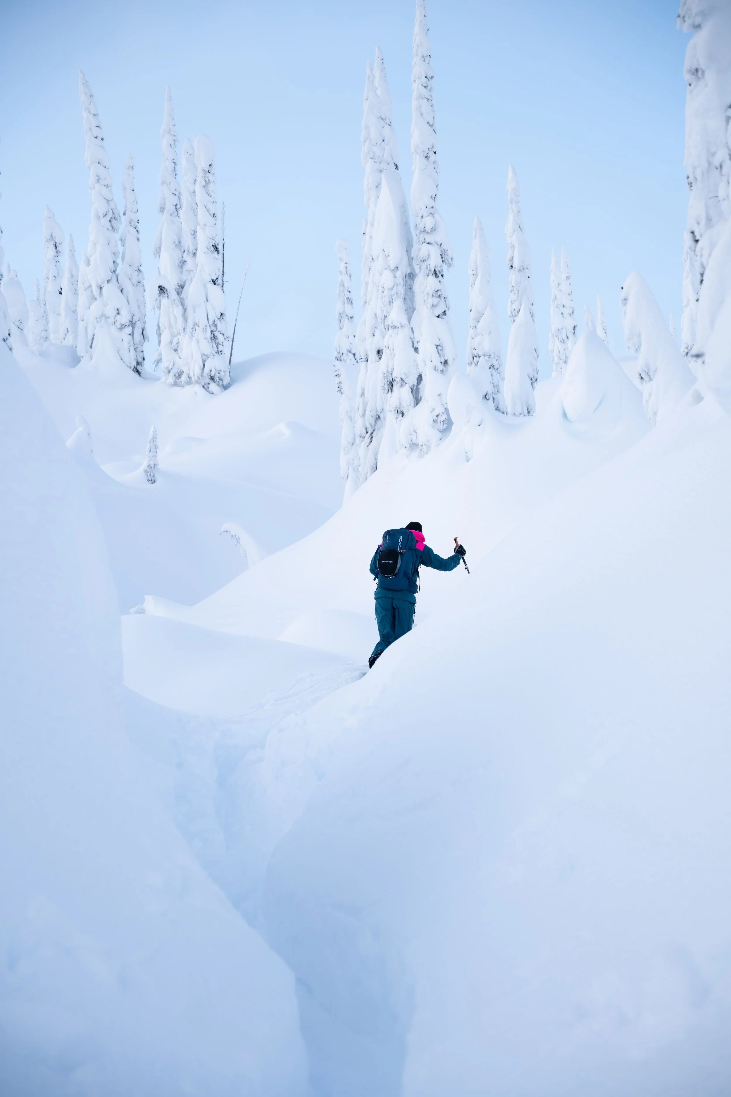 A person with a backpack hiking through deep snow in a snowy forest with tall, snow-covered evergreen trees under a partly cloudy sky.