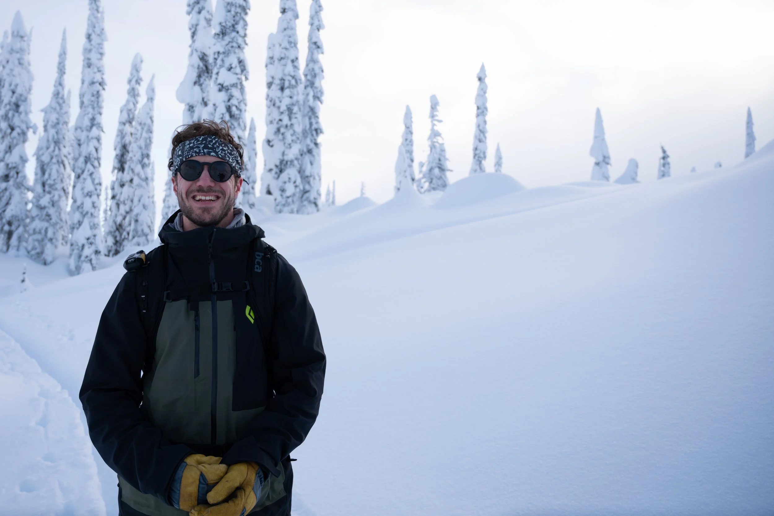 A man wearing sunglasses, a patterned headband, a black jacket, and yellow gloves stands in a snowy landscape with snow-covered trees in the background.