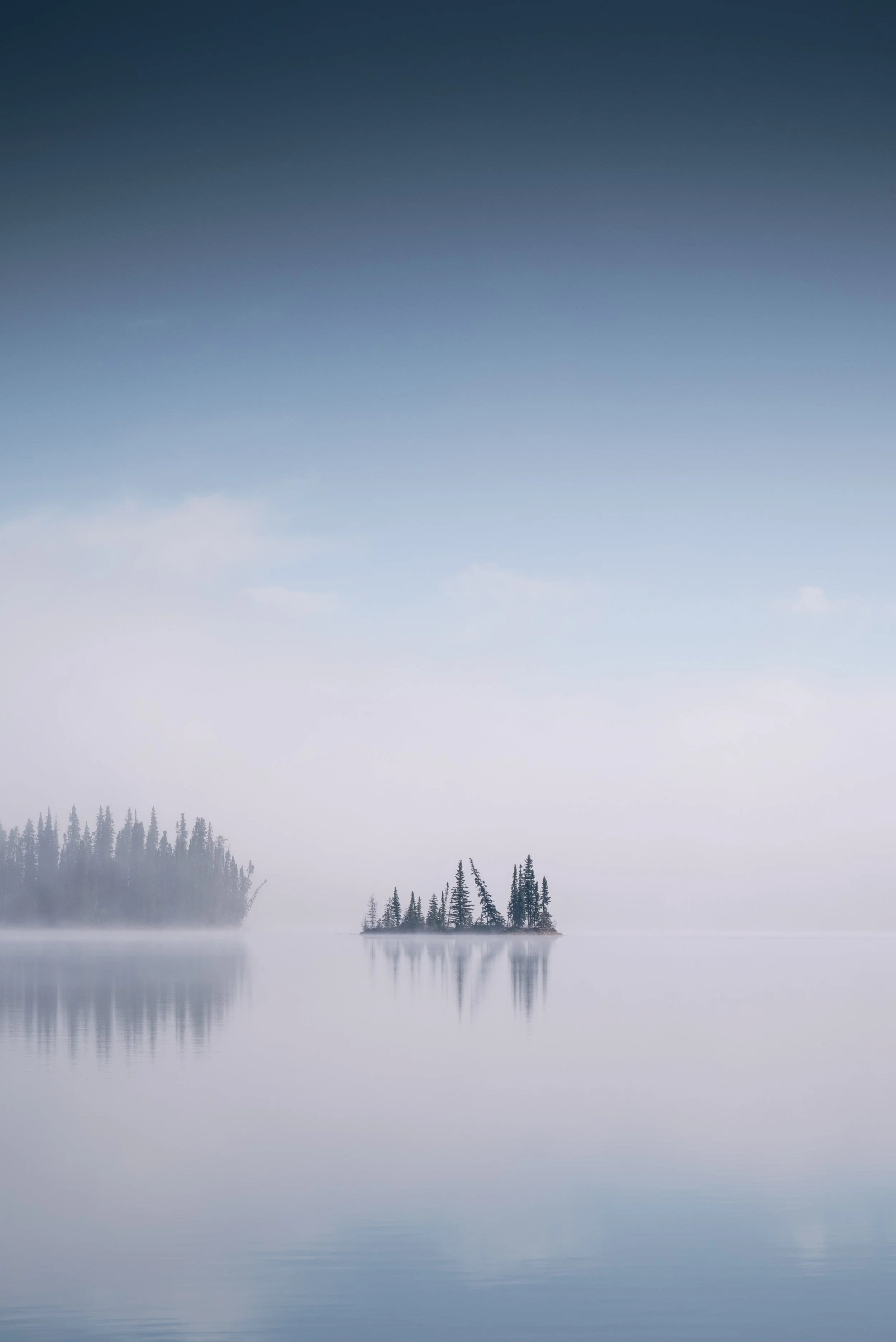 A calm lake with a small island of pine trees, surrounded by fog and a gradient sky from dark blue to light blue.