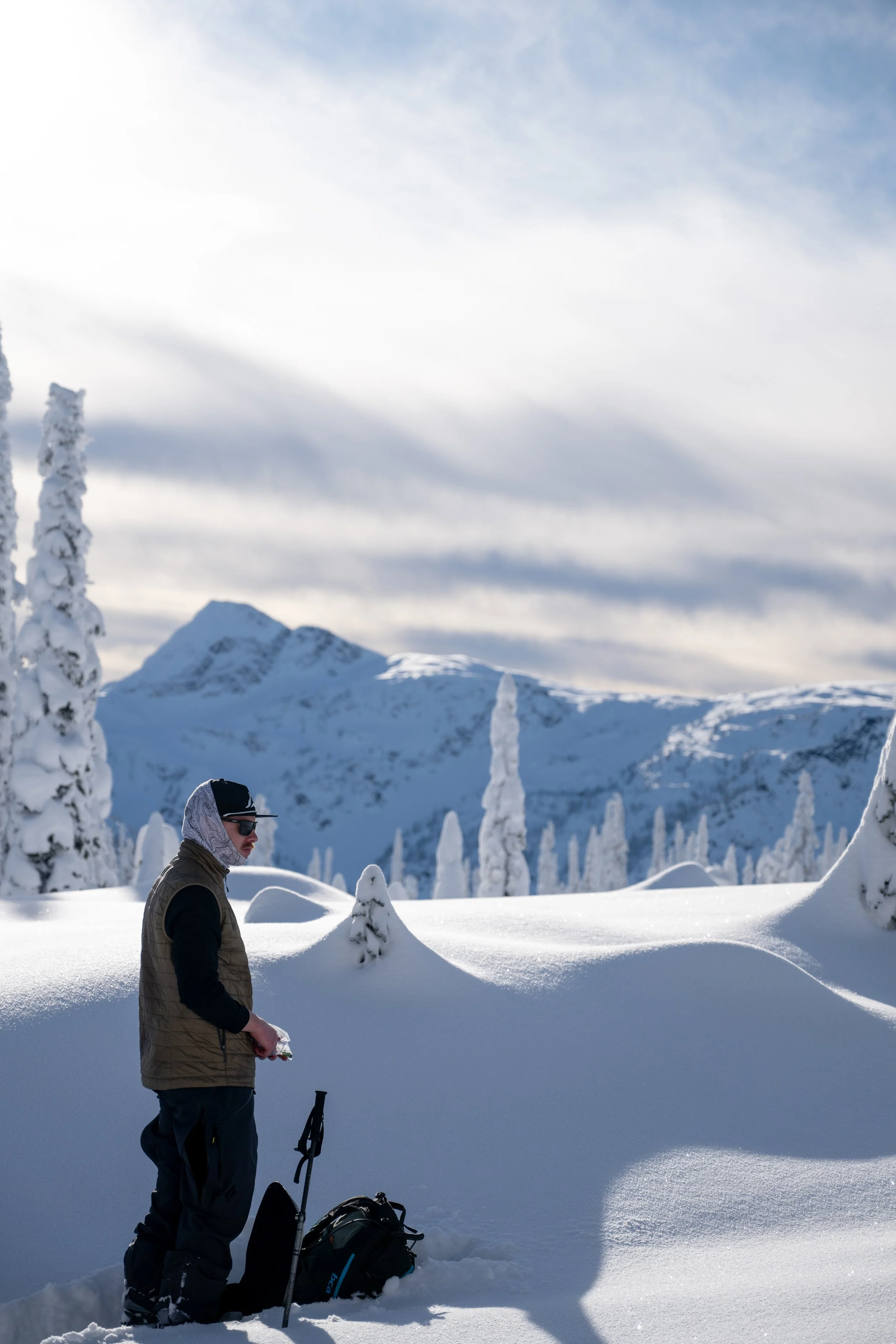 Person in winter clothing standing in a snowy landscape with snow-covered trees and mountains in the background.