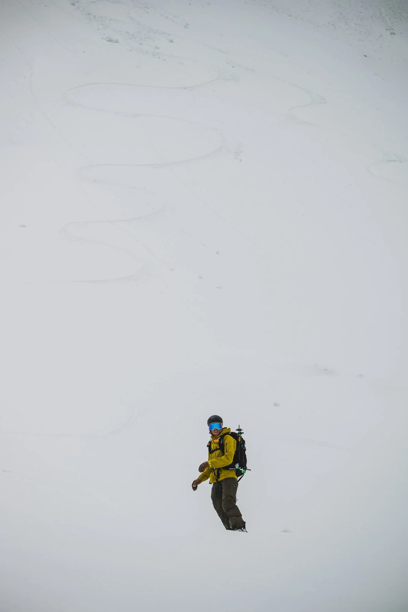 A person in winter gear standing in deep snow, wearing a yellow jacket, brown pants, a helmet, and skiing goggles, with ski equipment on their back. Montana backcountry skiing