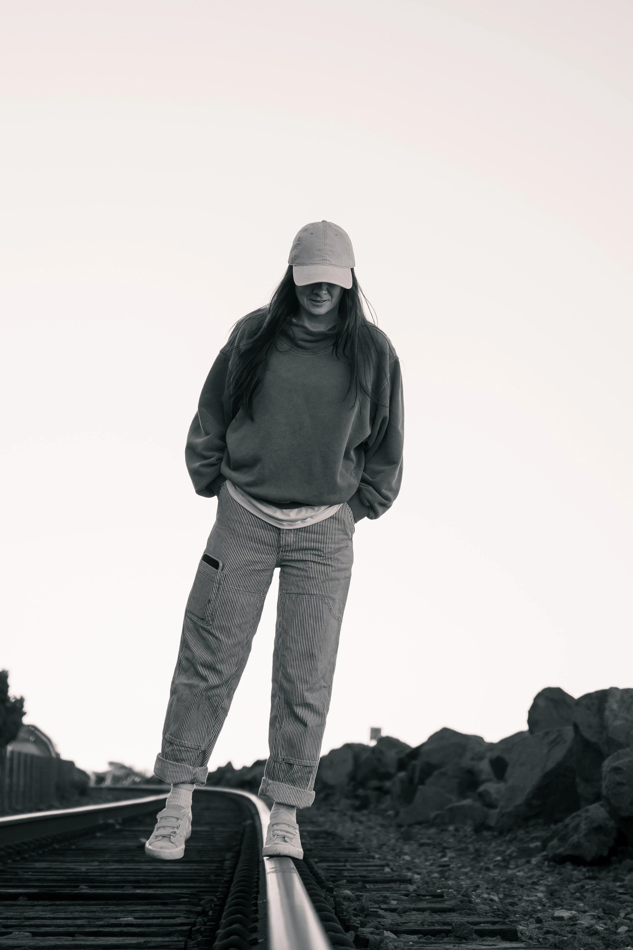 A woman in casual clothing and a baseball cap standing on railway tracks outdoors in black and white.