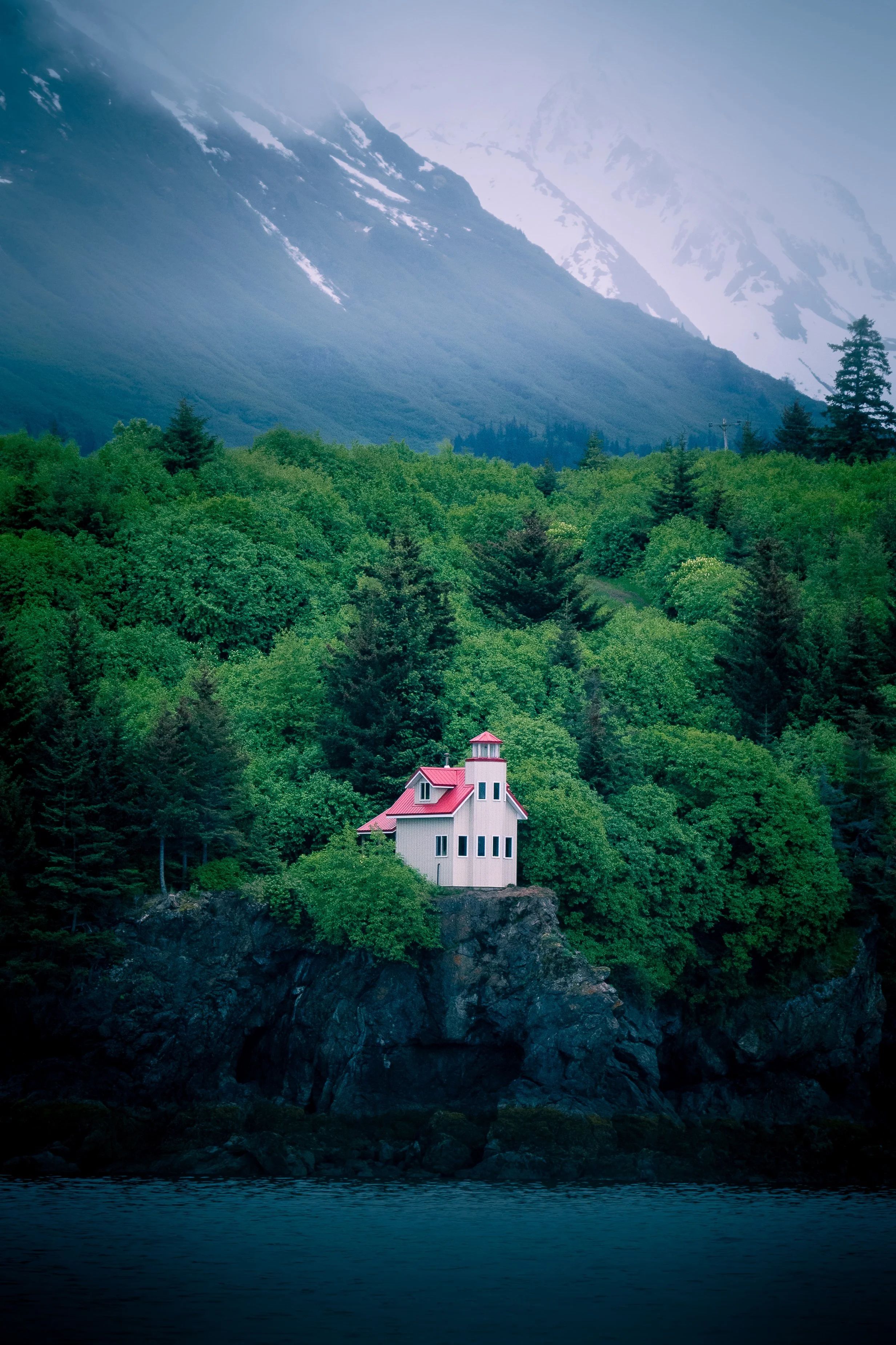 A house built on a rocky coastline surrounded by dense green forest and tall mountains in the background.