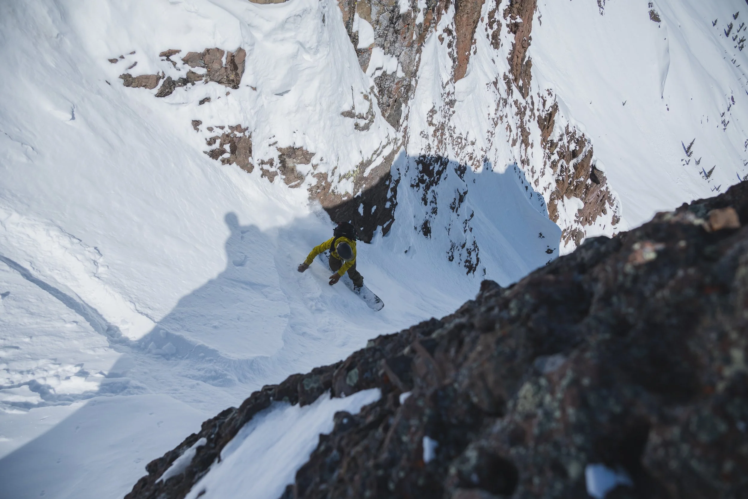 A skier wearing a yellow jacket and black helmet skiing down a steep snow-covered mountain slope with rocky sections, viewed from above.