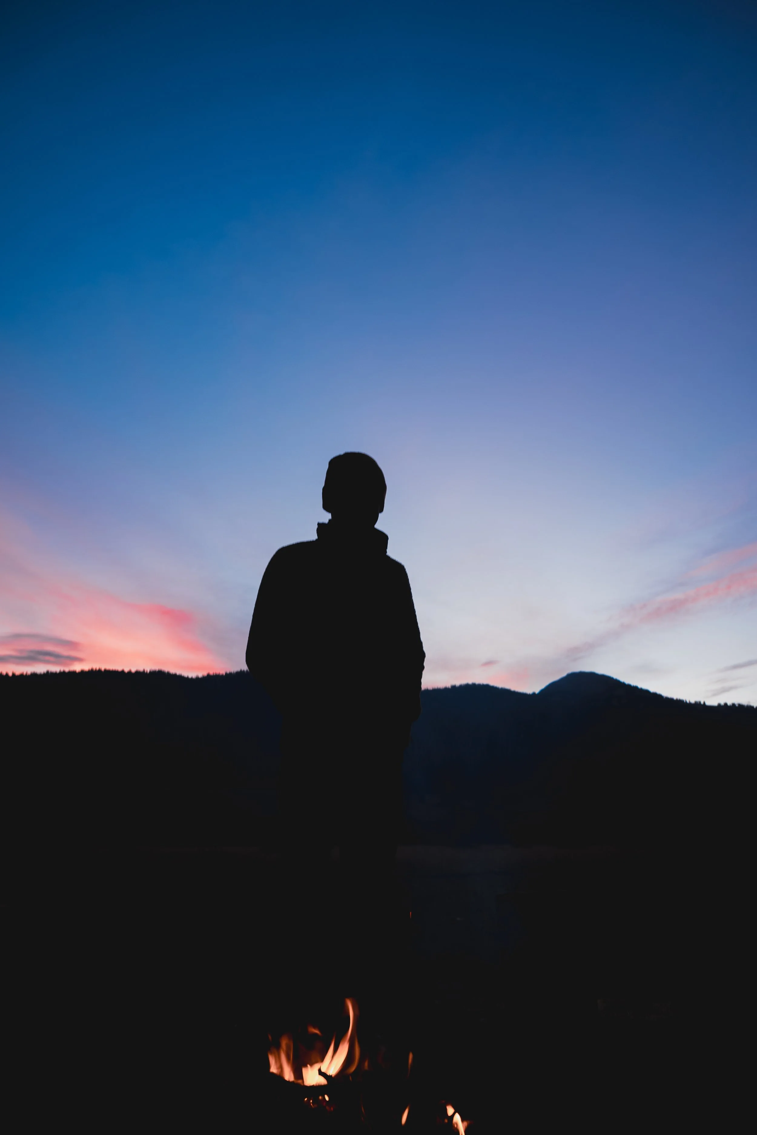 Silhouette of a person standing outdoors at sunset, with mountains in the background and a campfire burning in the foreground.