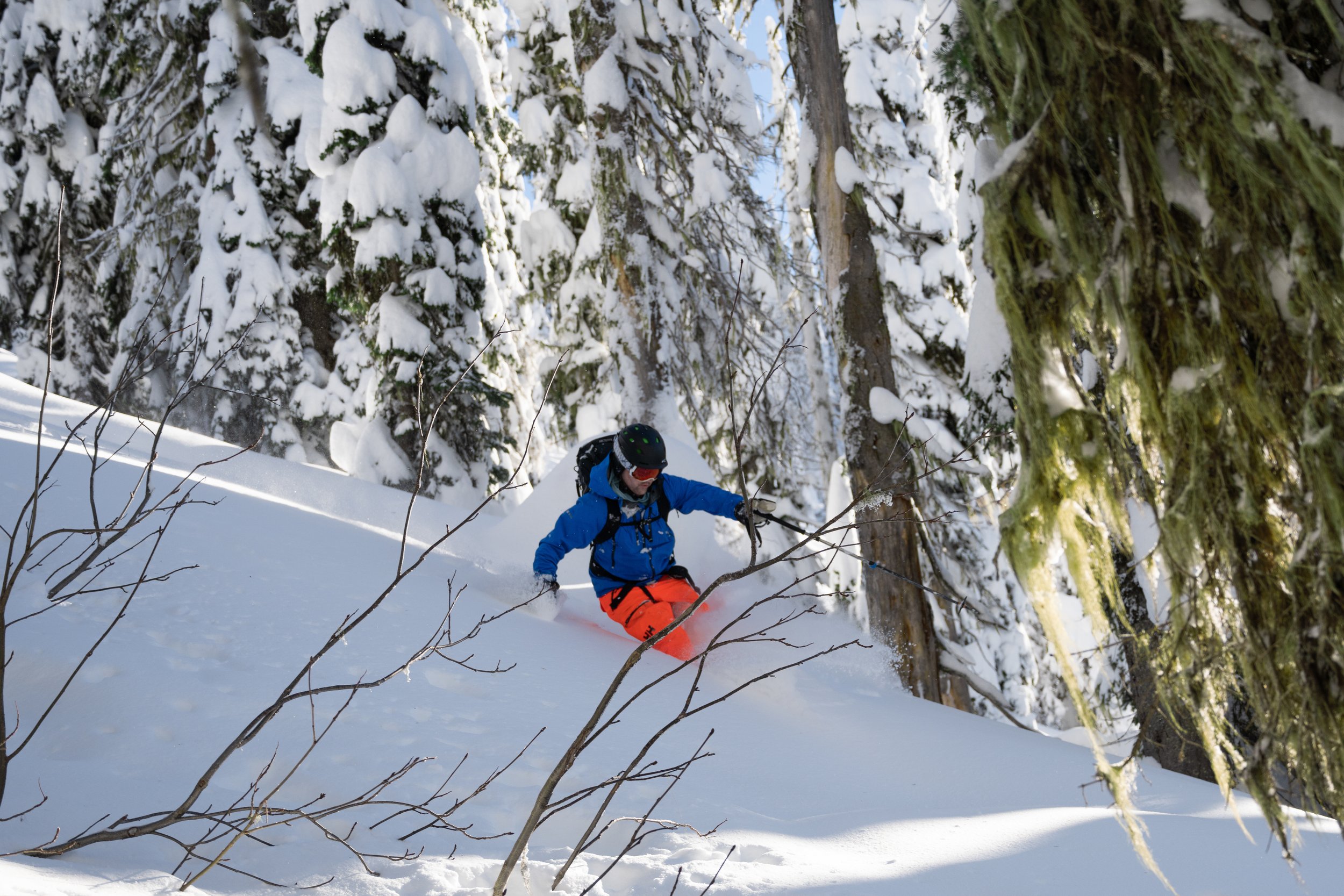 A person snowboarding in a snowy forest, wearing a blue jacket, orange pants, a black helmet, and goggles, surrounded by snow-covered trees.