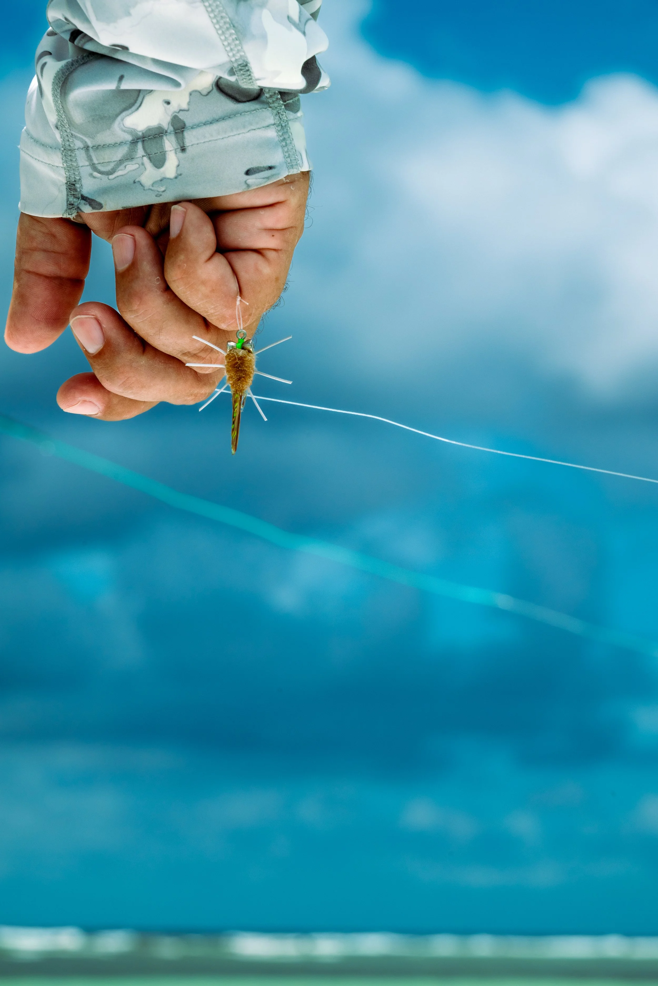 A person in camouflage clothing holding a fishing lure resembling a small fish with feathers, against a background of a cloudy sky.