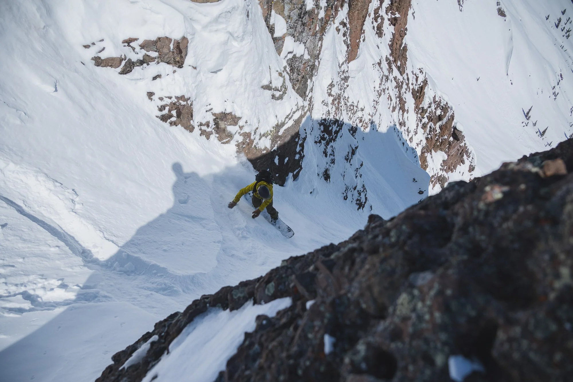 A person in yellow gear snowboarding down a steep, snowy mountain slope with rocky cliffs around. montana backcountry skiing